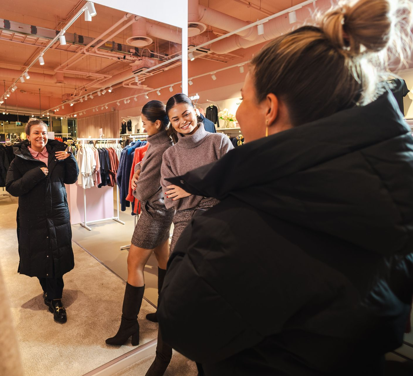 Three women in a clothing store, one taking a photo of two others. Soft pink walls, racks of clothes.