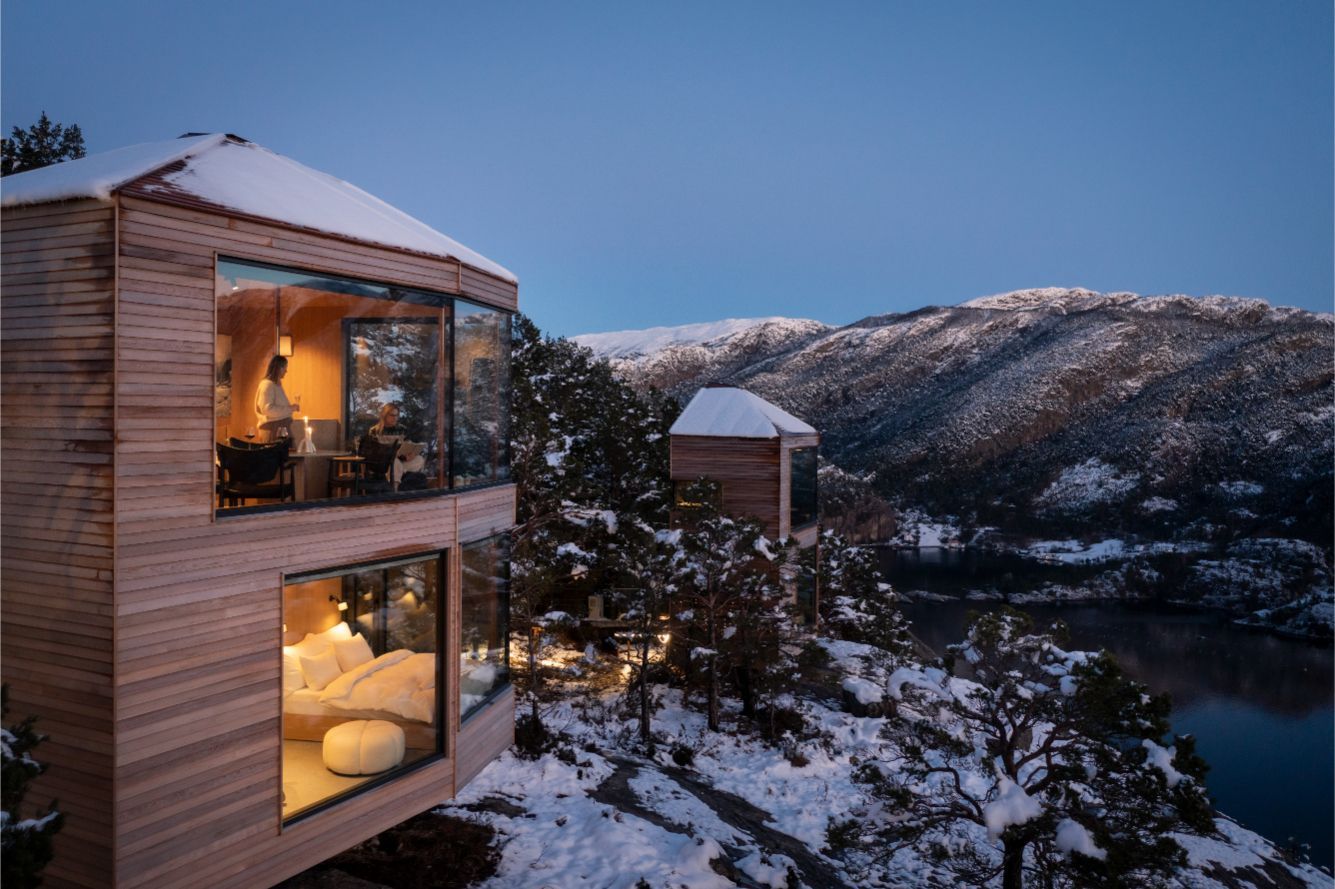 Wooden cabin with large windows overlooking a snow-covered landscape and lake at dusk.