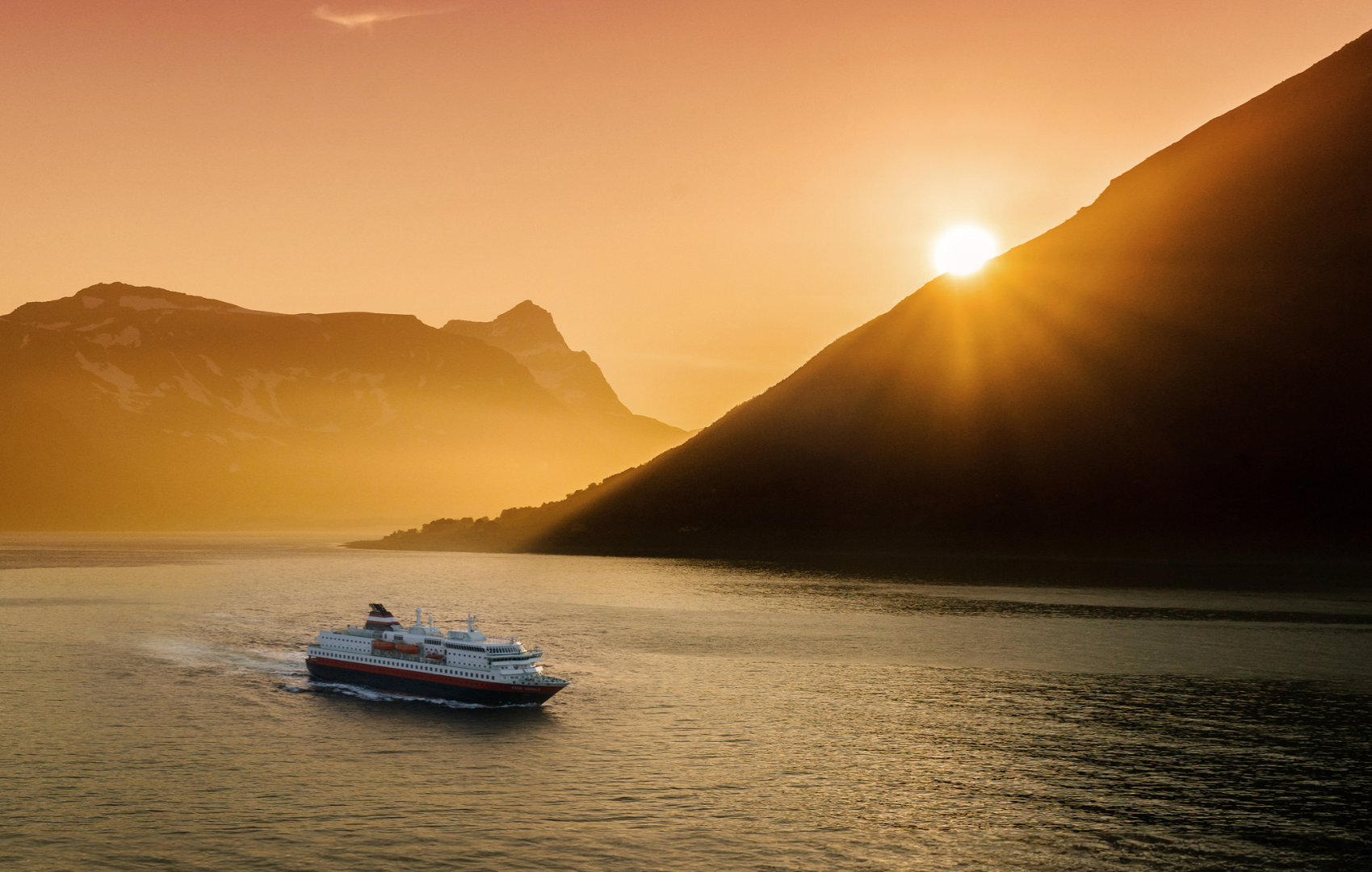 Ferry sailing through a fjord, with mountains in the background during a sunset.