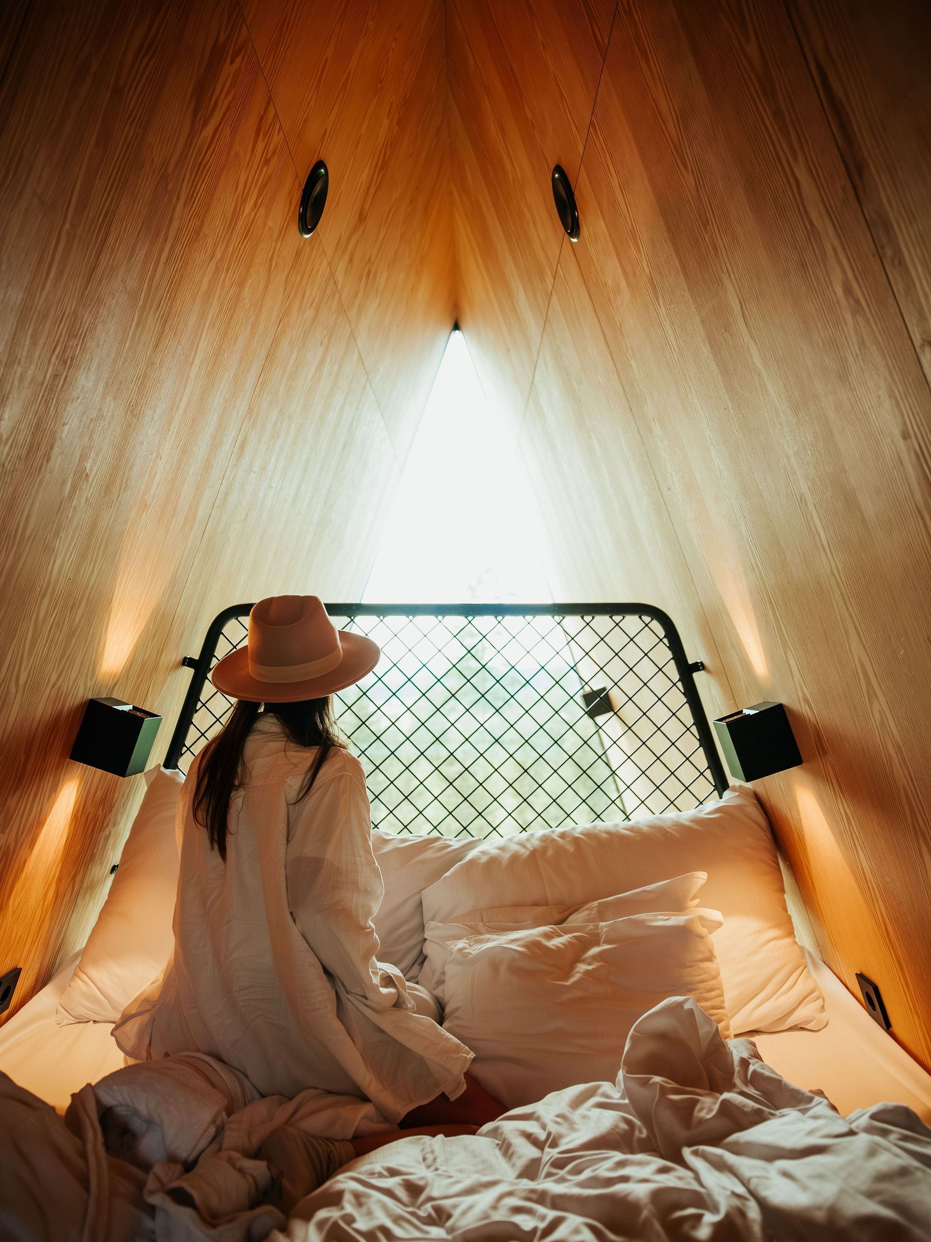Woman sitting on a bed inside a tent, wearing a hat, looking out a window.