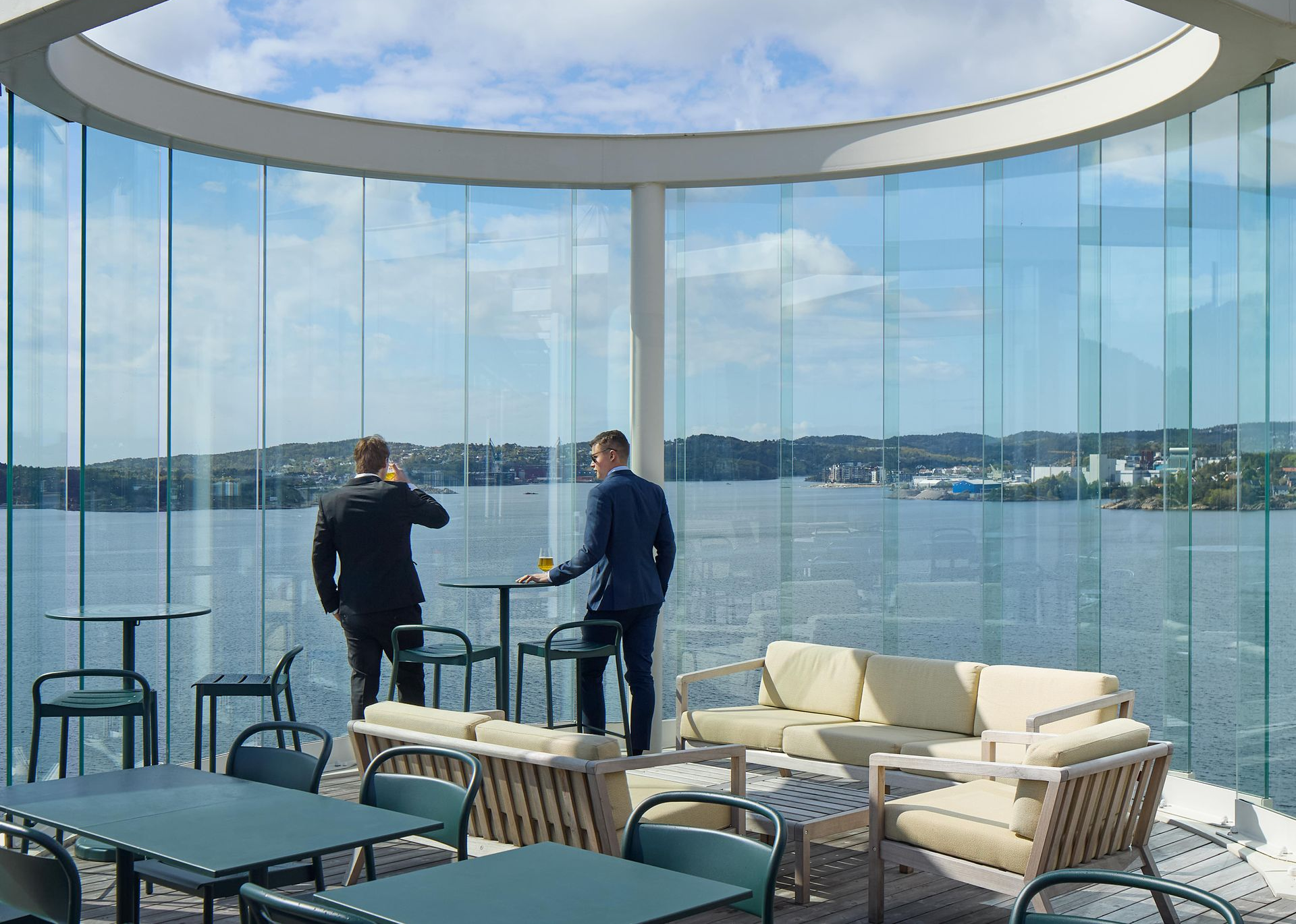 Two people standing at a table on a waterfront patio. Curved glass walls, blue sky, water view.