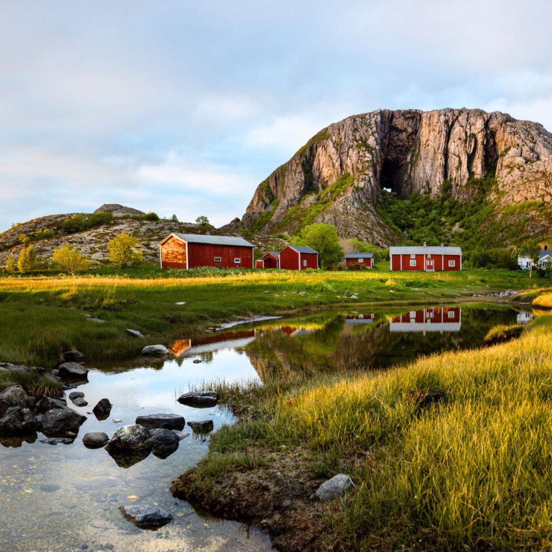 Red buildings reflected in a calm pond, with a large rock formation in the background under a blue sky.