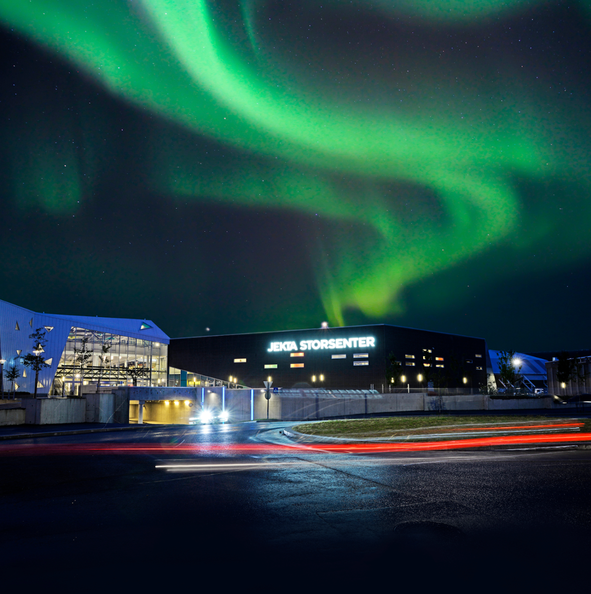 Aurora borealis over Jevnaker Storsenter, Norway.  Buildings are lit, car lights streak across the road. Green lights dance in the sky.