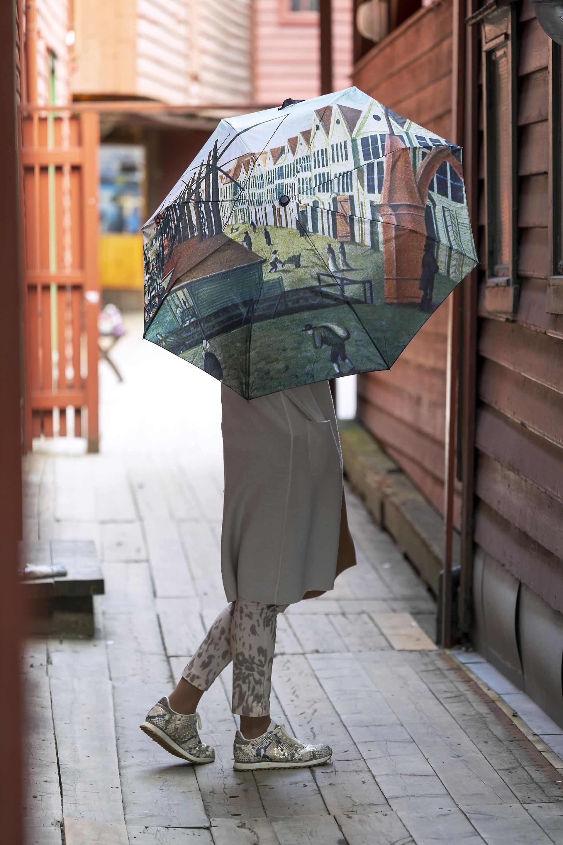Person walking under an umbrella featuring a cityscape painting; walking on a wooden walkway.