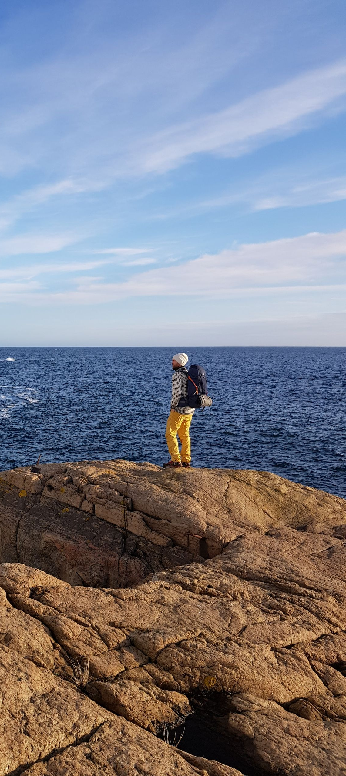 Person with backpack stands on rocky shore, gazing at the ocean under a blue sky.