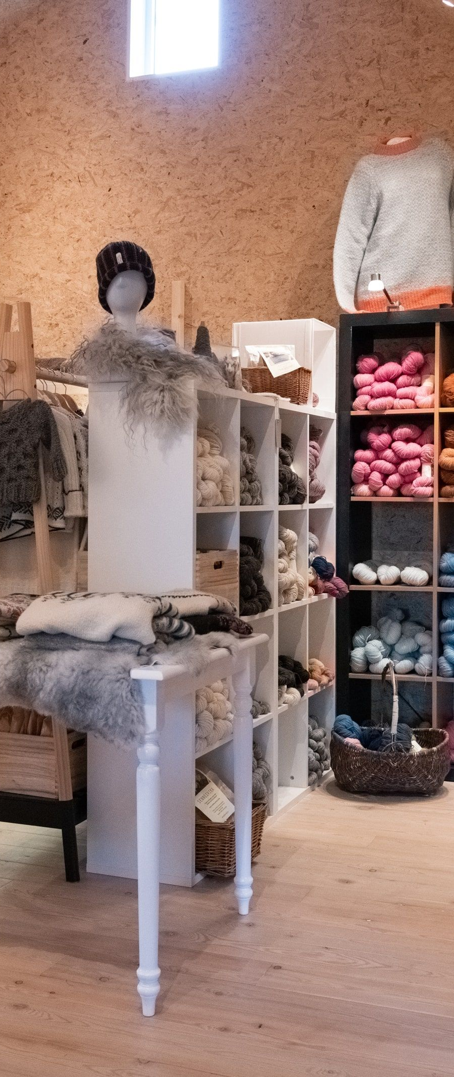 A brightly lit retail shop displays various yarn and clothing items on white shelves and a table.