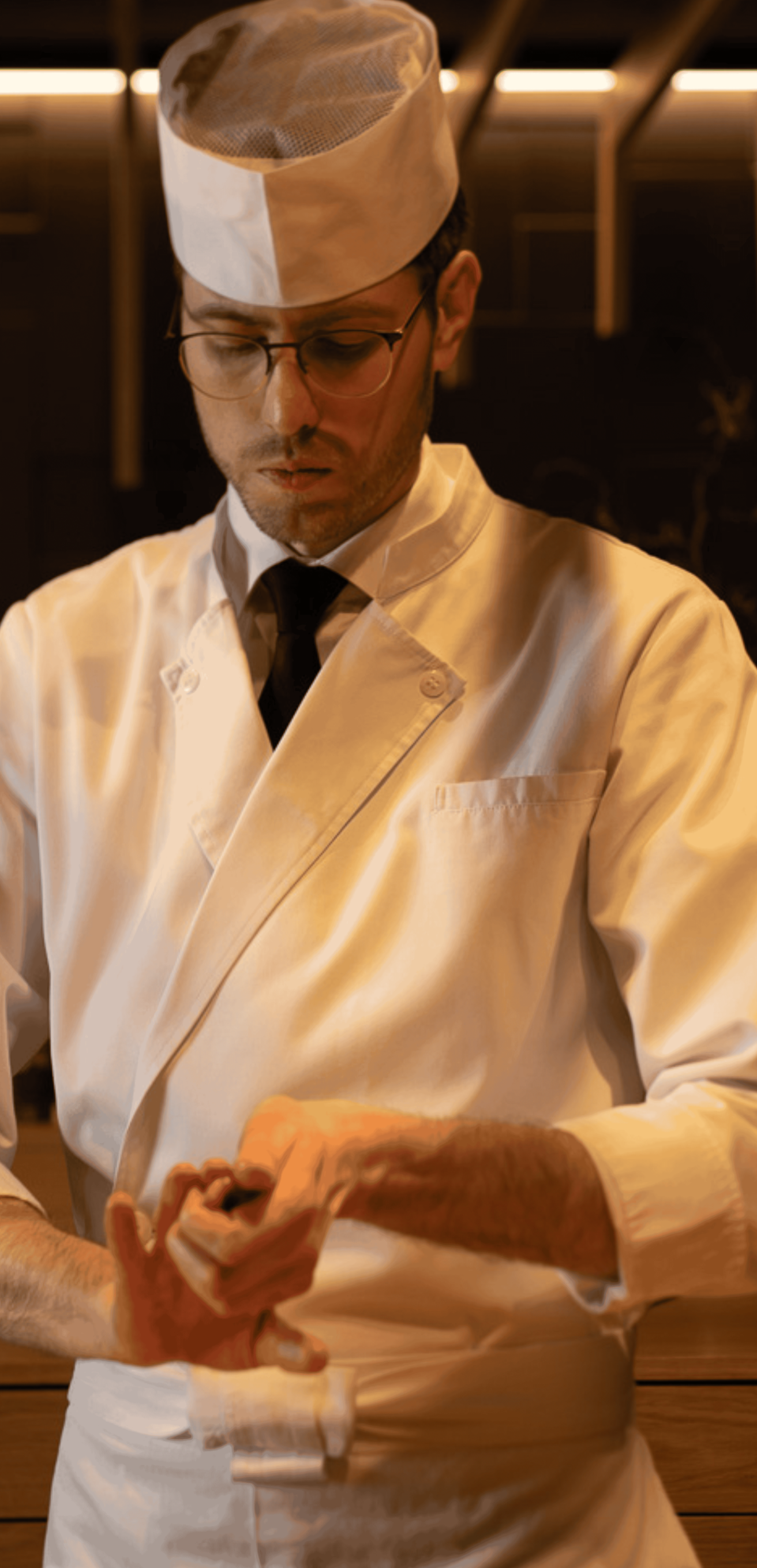 Sushi chef preparing food. He wears a white uniform and hat, with a focused expression.