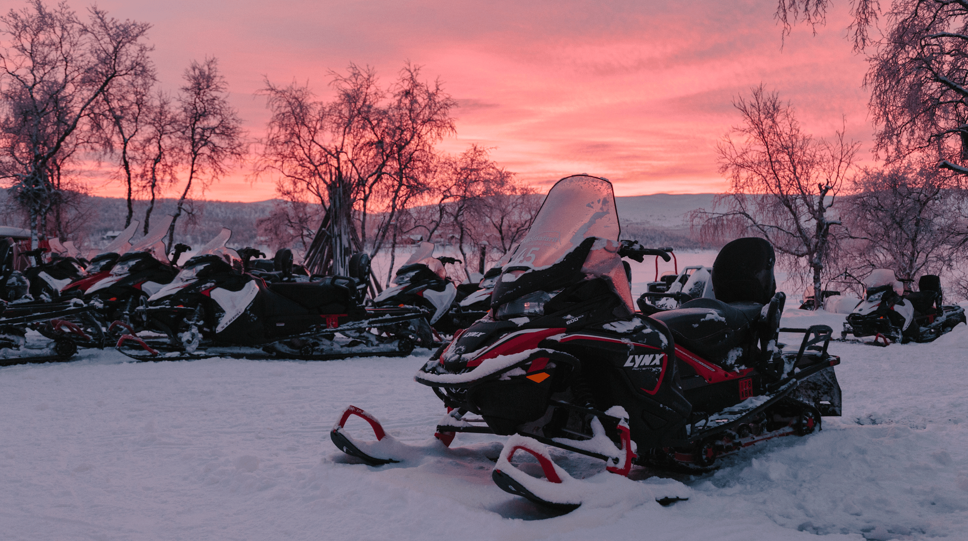 Snowmobiles parked in a snowy landscape, under a pink and orange sunset.
