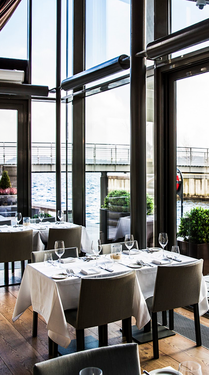 Restaurant interior with tables set for dining, overlooking water. Large windows, neutral color palette.