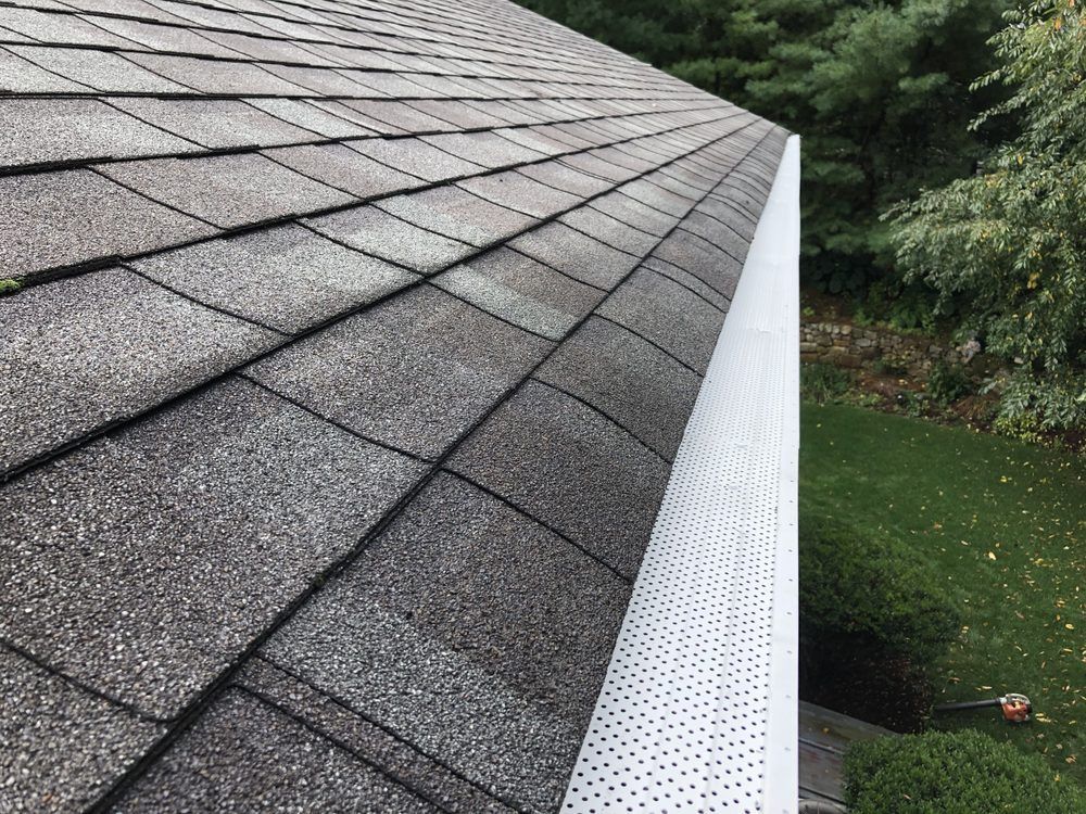 Close-up of a roof with gray asphalt shingles and a white gutter with small holes, viewed from above.