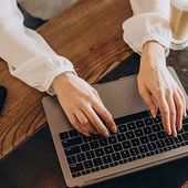 Hands typing on a laptop keyboard on a wooden table next to a glass of iced coffee.