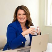 A smiling person in a royal blue blazer sits at a desk with a laptop, holding a white mug.