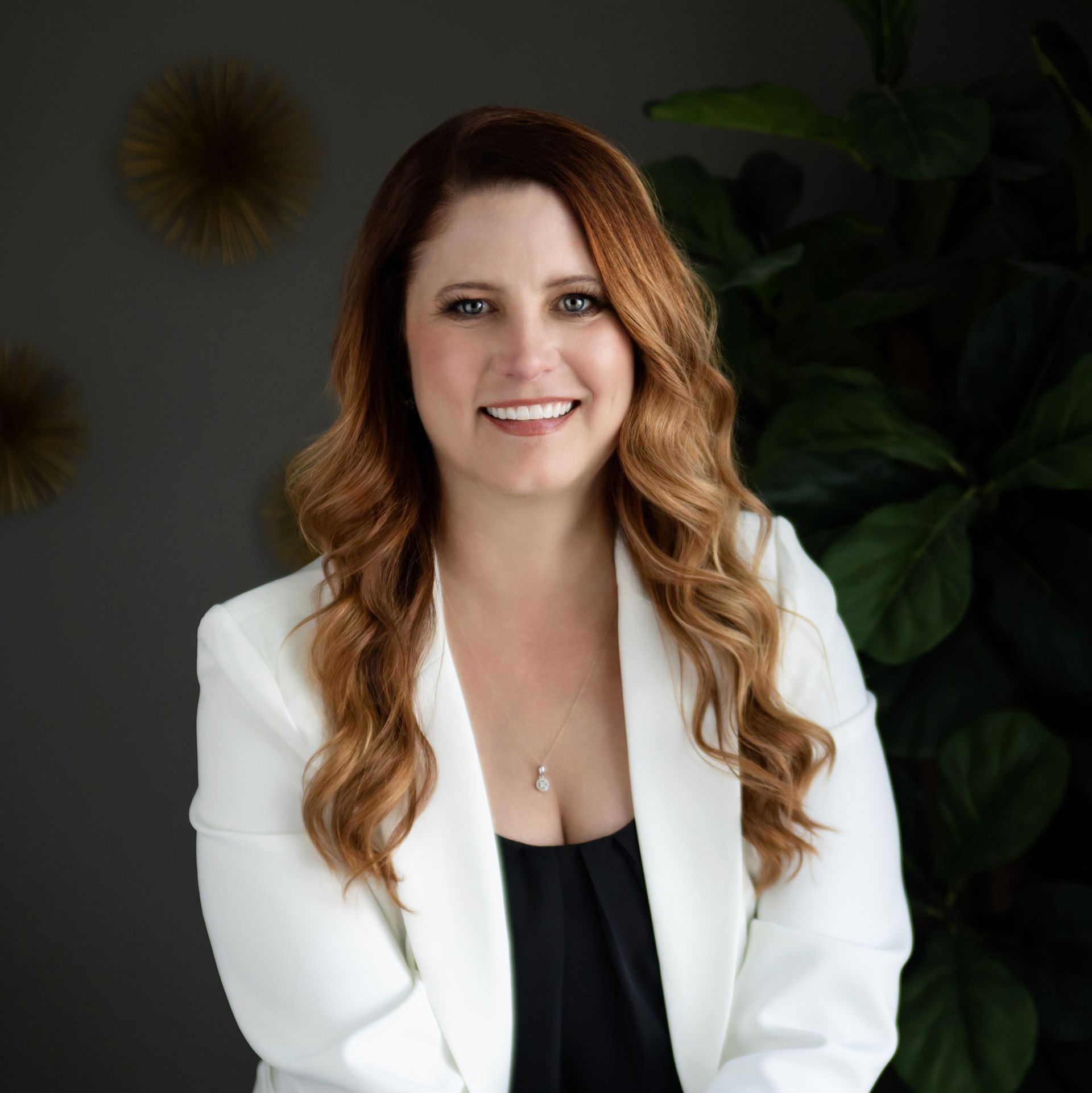 A person with long brown hair, wearing a black blazer and white top, smiles with arms crossed against a neutral wall.