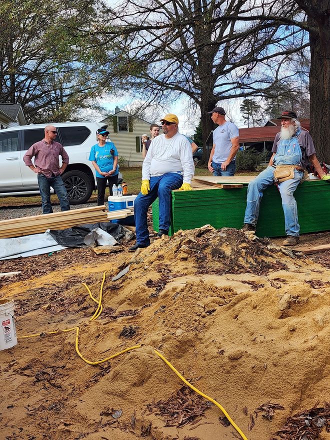 A group of people working on a construction project outdoors, surrounded by wood, dirt, and tools. The sky is overcast.