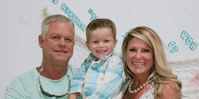 A family of three smiling at the camera. A man on the left, a young boy in the middle, and a woman on the right. They are indoors.