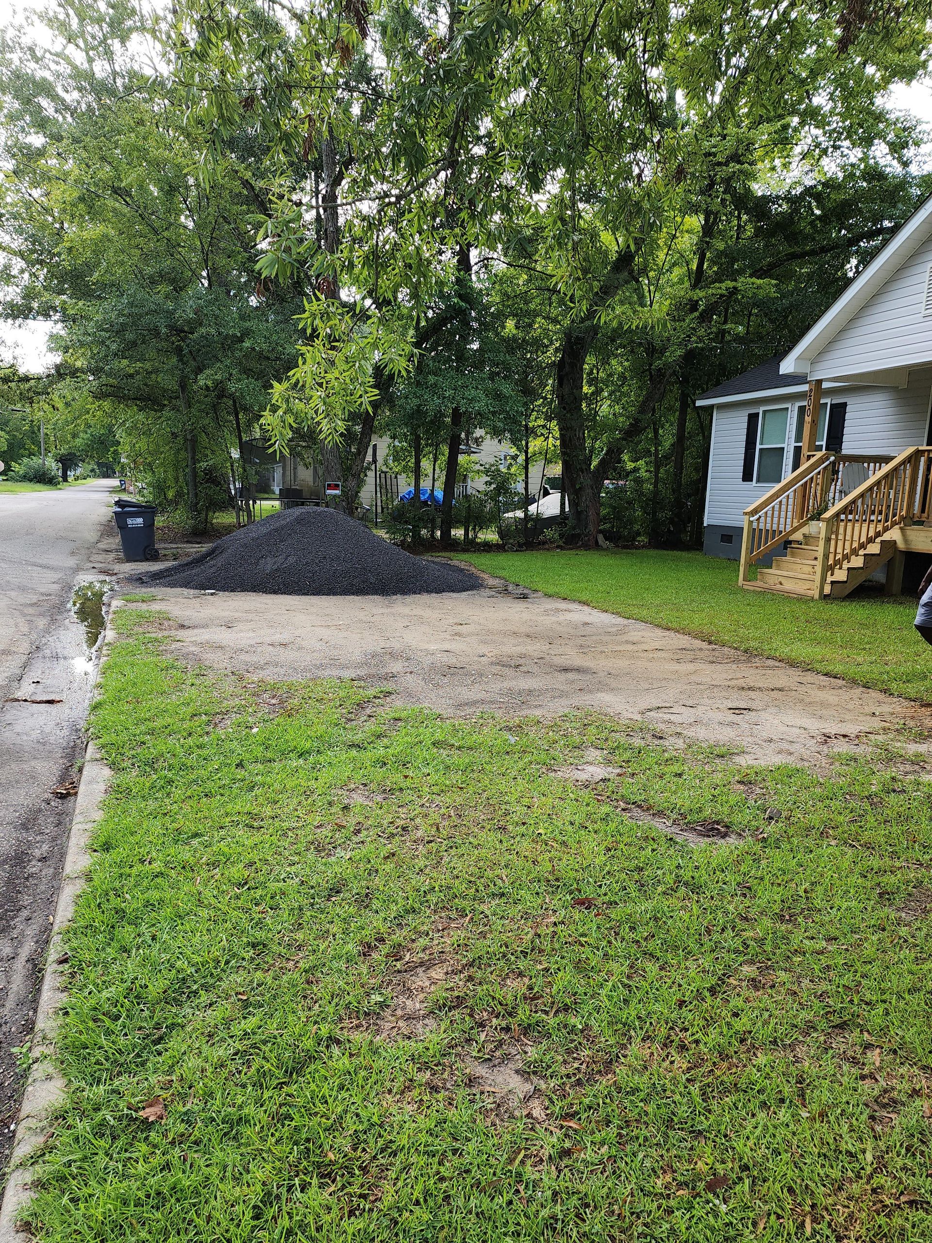 A pile of dark material sits on a dirt patch beside a house with wooden steps, next to a sidewalk and grass.