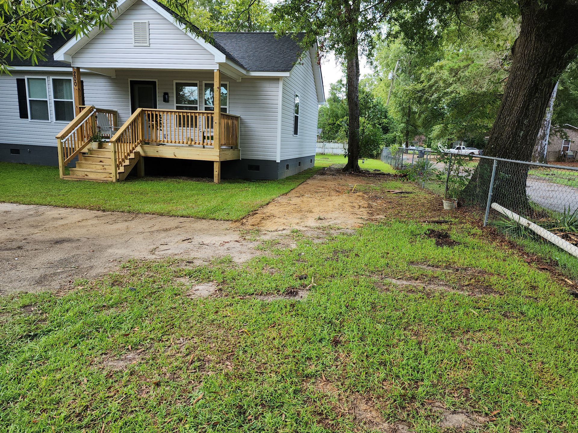 Small, light-colored house with wooden porch and steps. Partially overgrown yard with dirt driveway.