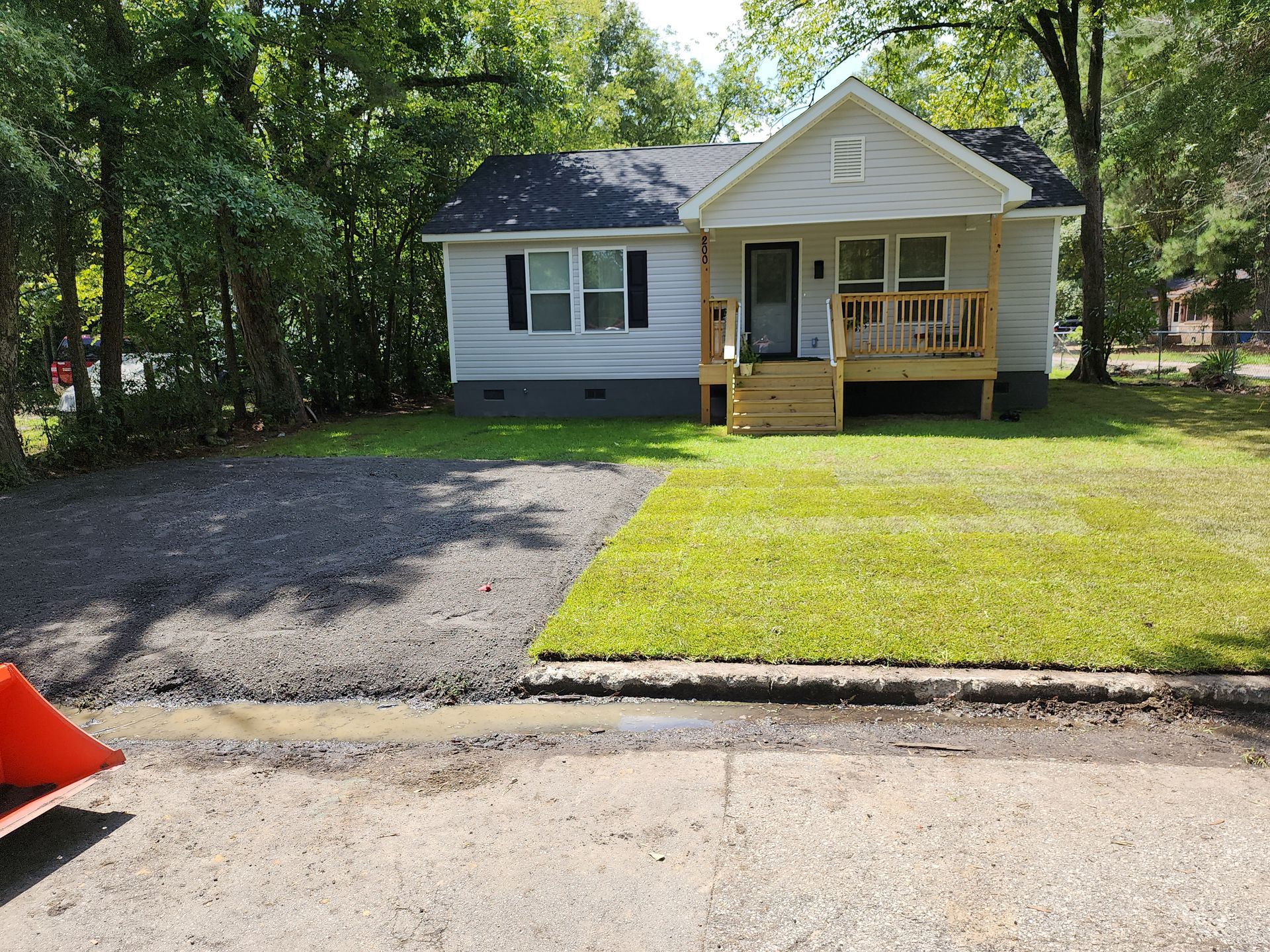 Small gray house with a porch and grass lawn, beside a newly paved driveway. Trees in the background.