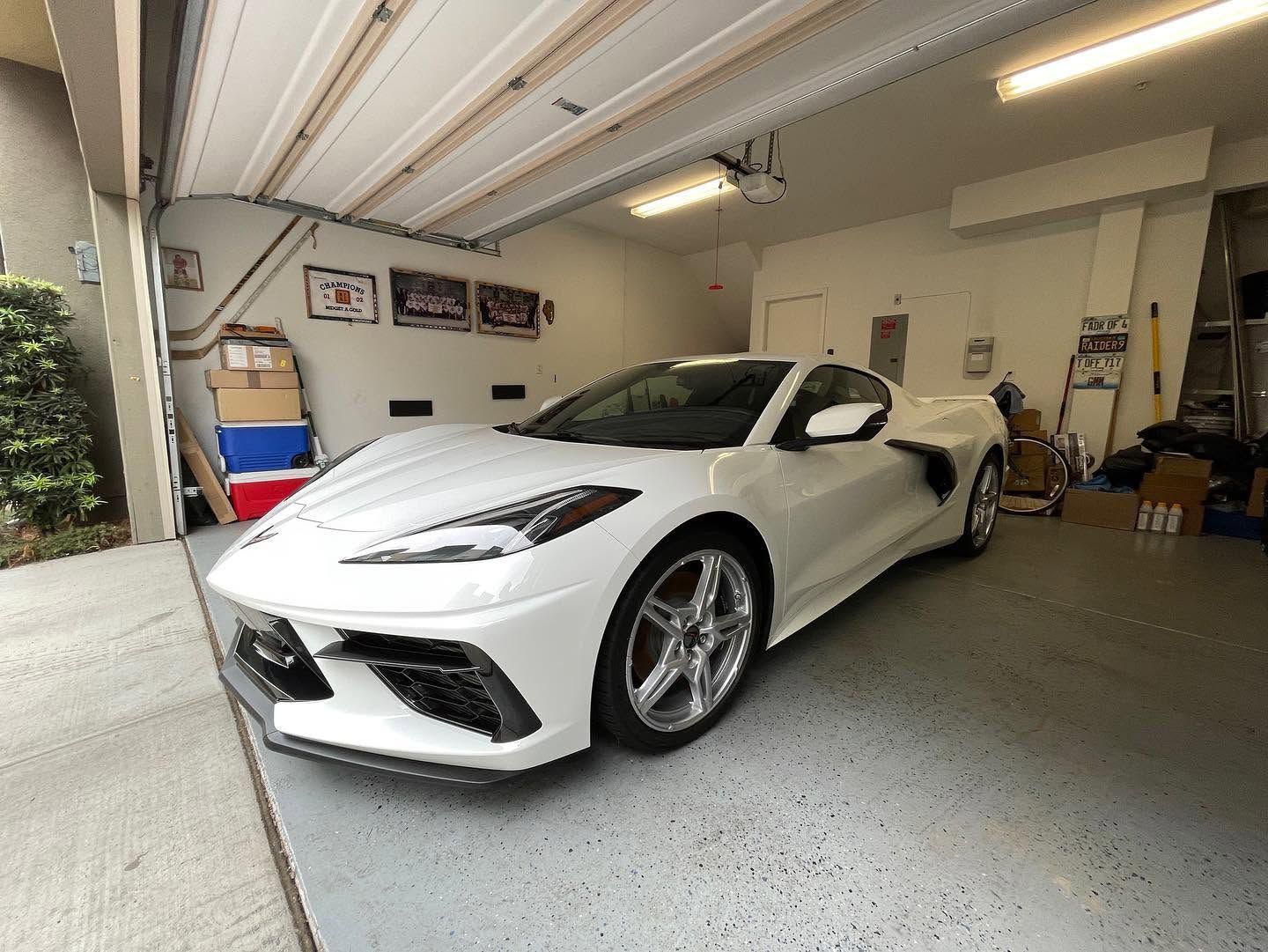 White sports car parked in a garage with open door, shelves, and tools around it.