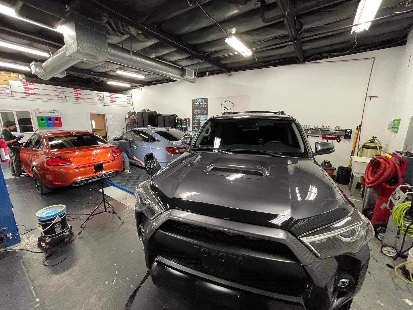 Auto repair shop with a black SUV in front, an orange sports car, and a gray car in the background.