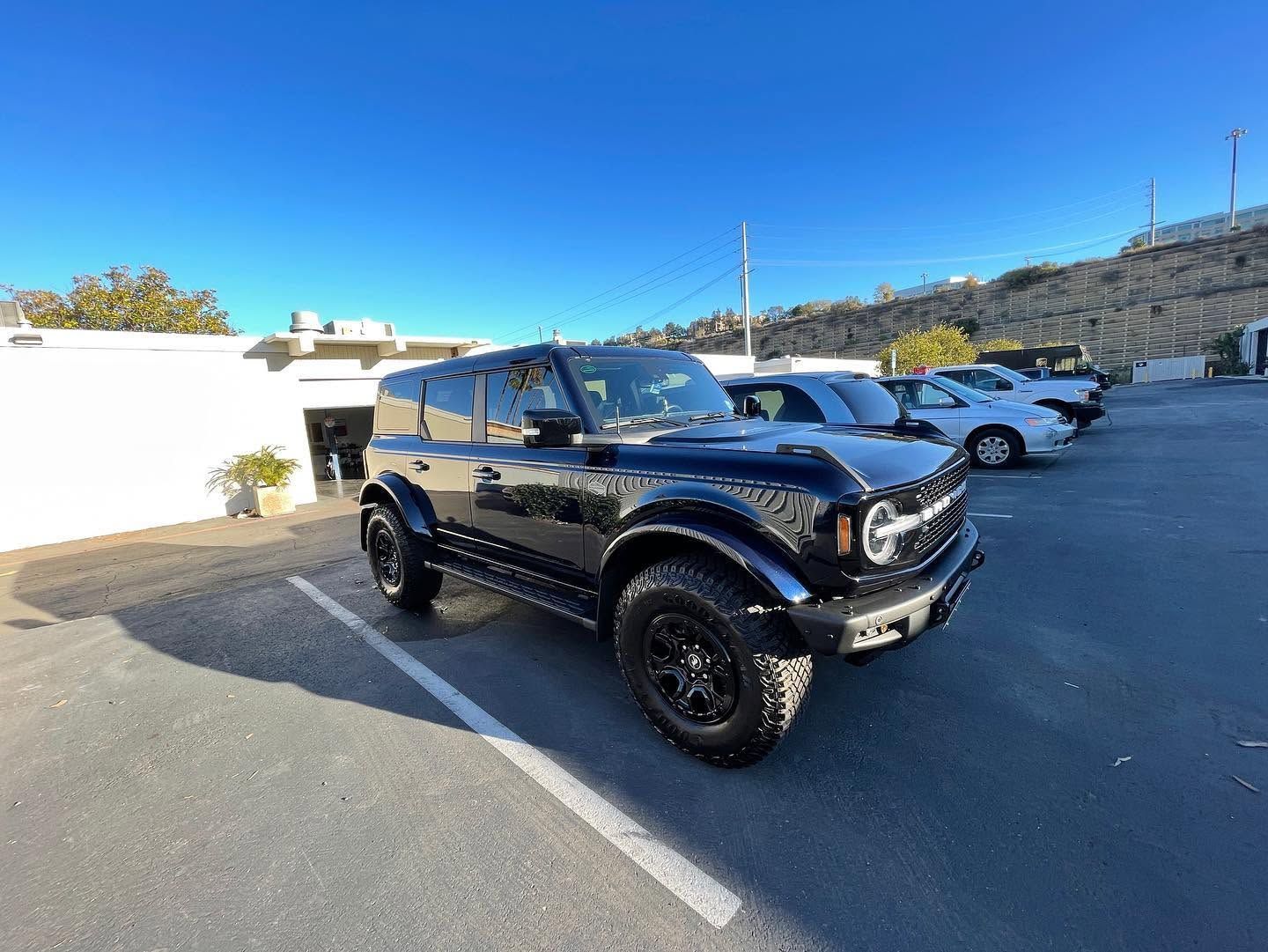 Black off-road SUV parked in a lot under a clear blue sky, with other cars and a hillside in the background