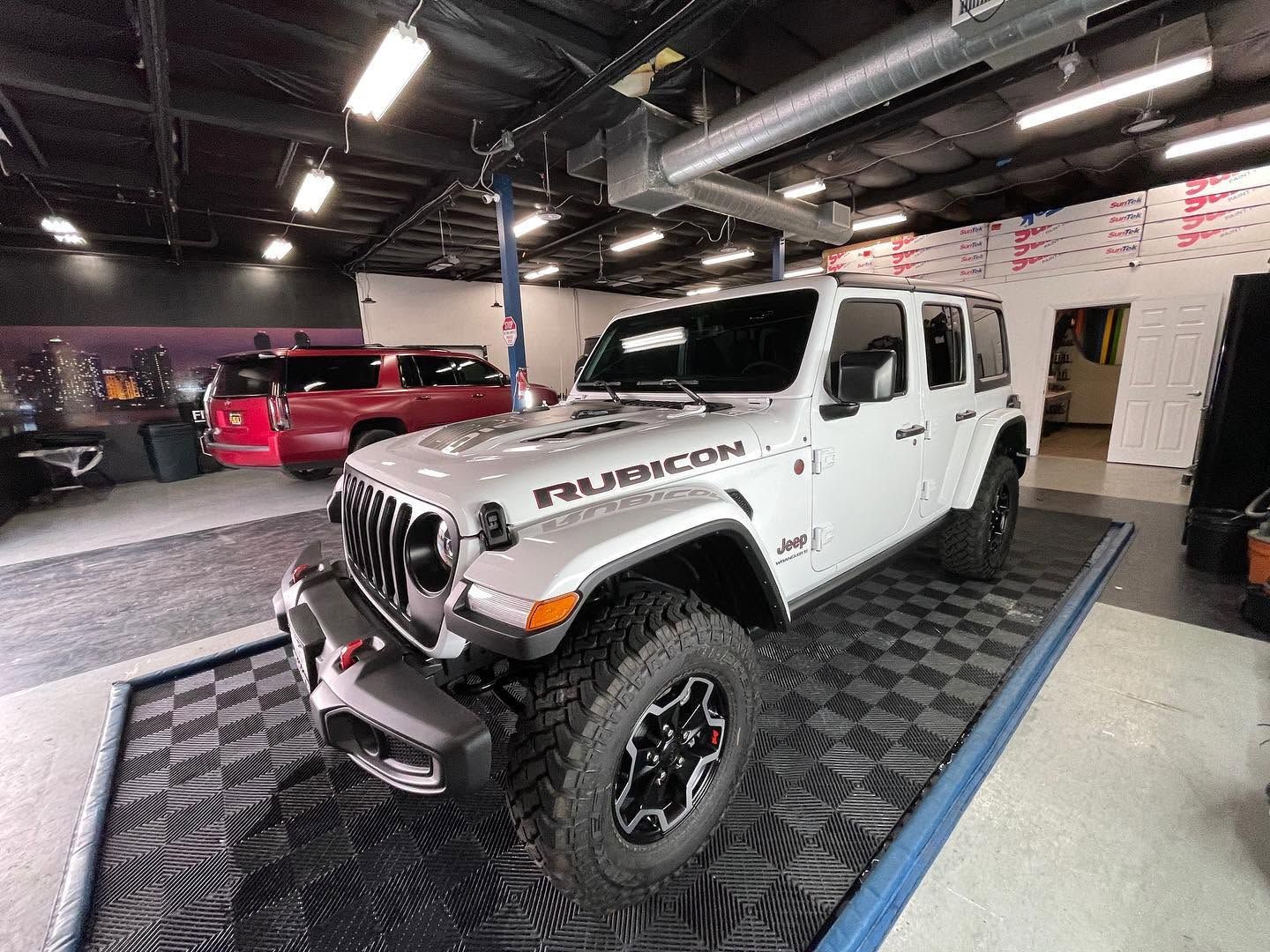 White Jeep Rubicon displayed indoors on a checkered platform in a showroom, with other vehicles behind it.