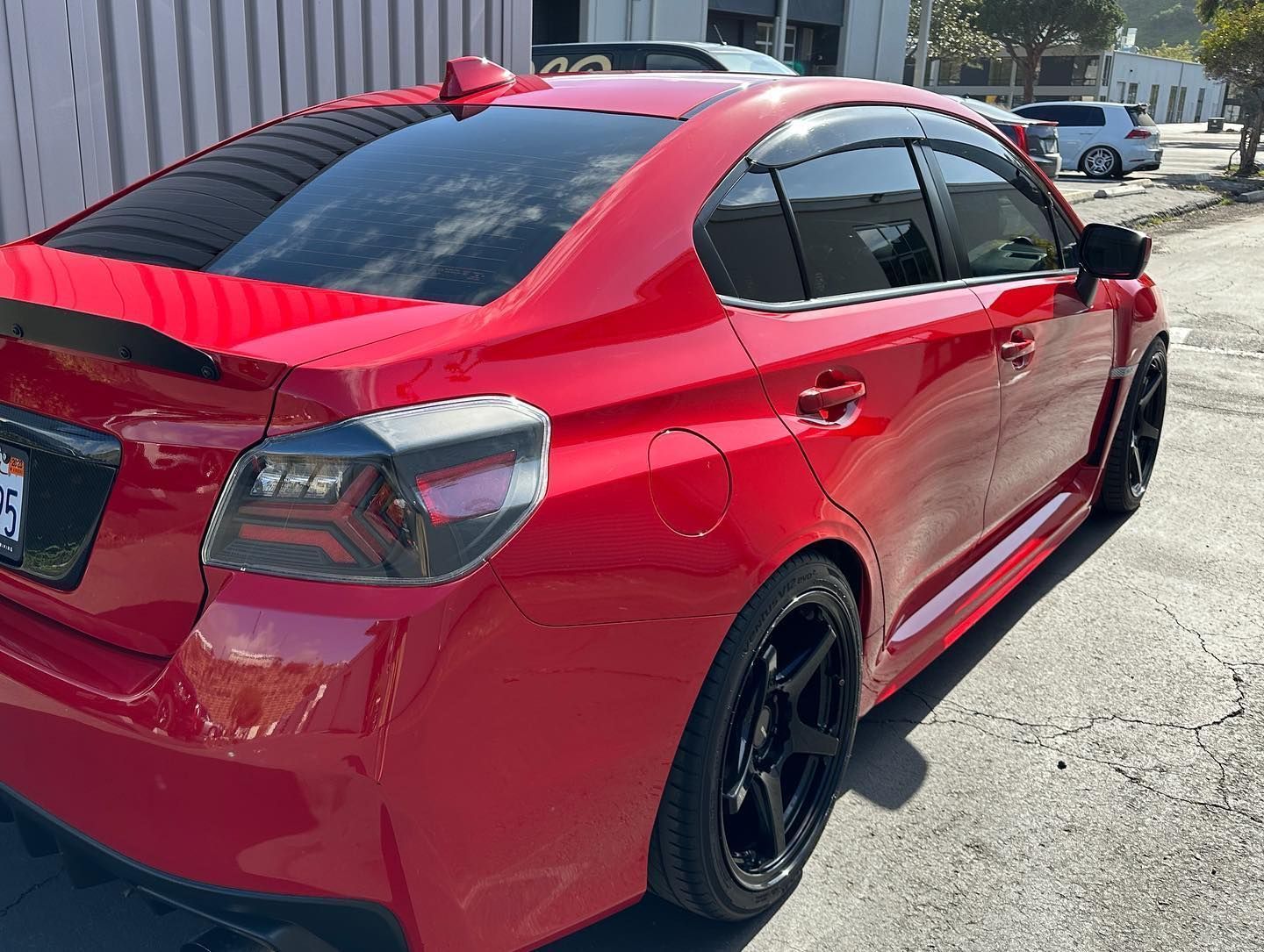 Red sedan parked beside a building on a sunny street, rear three-quarter view.