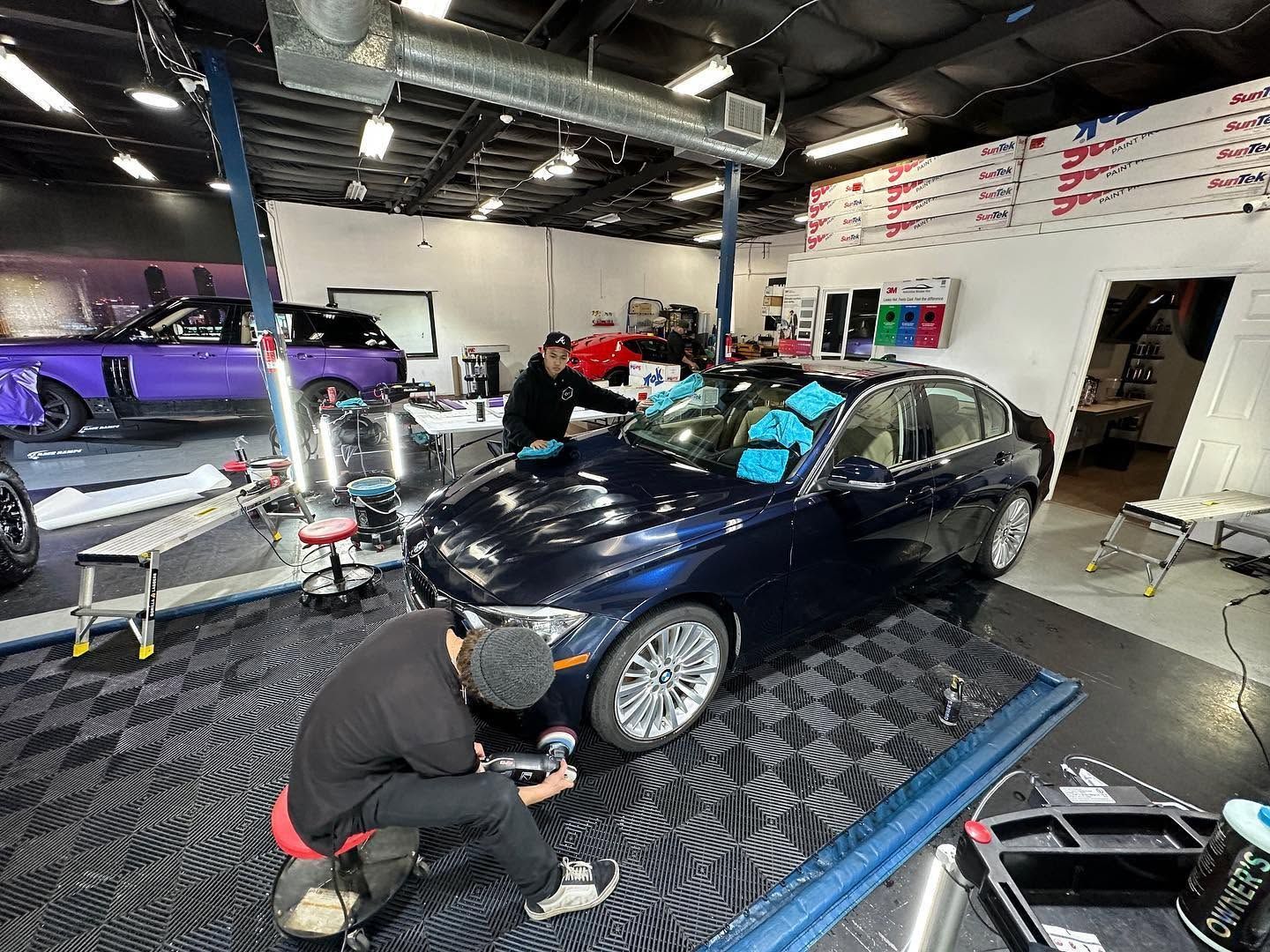 Mechanic polishing a black car in an auto detailing shop, with another technician working nearby.