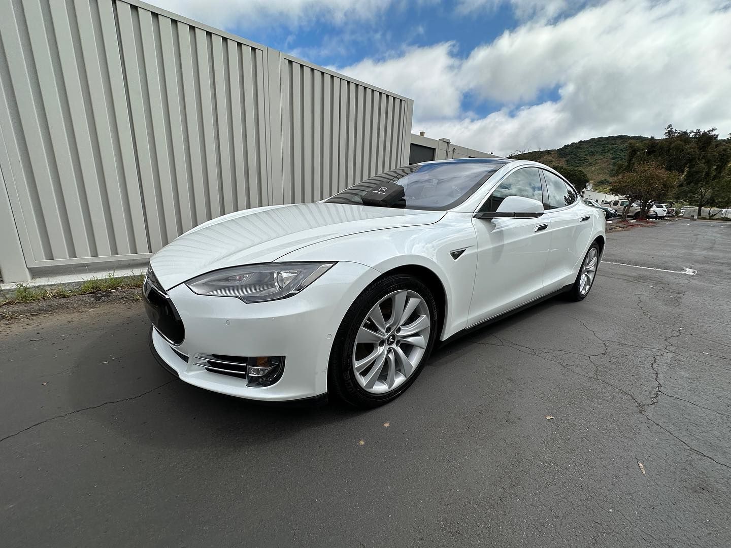 White Tesla sedan parked on wet pavement beside a large corrugated metal building under cloudy skies