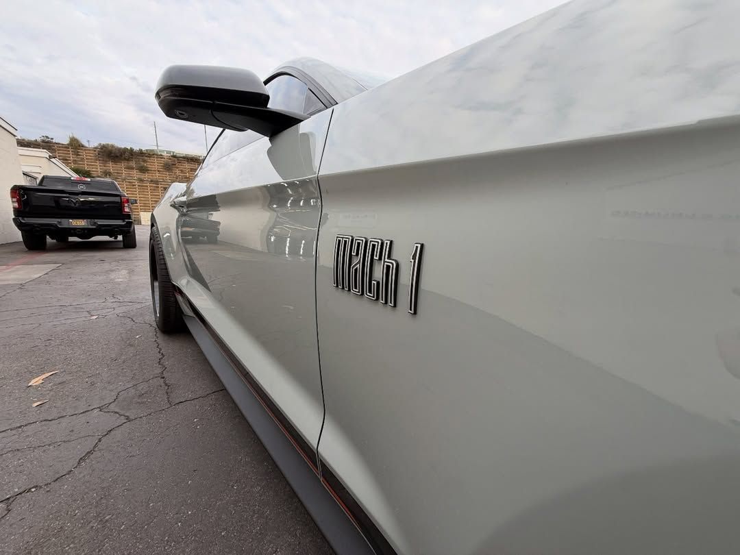 Close-up of a white Mustang badge on a car parked on a road with another car and rocky hills in the background