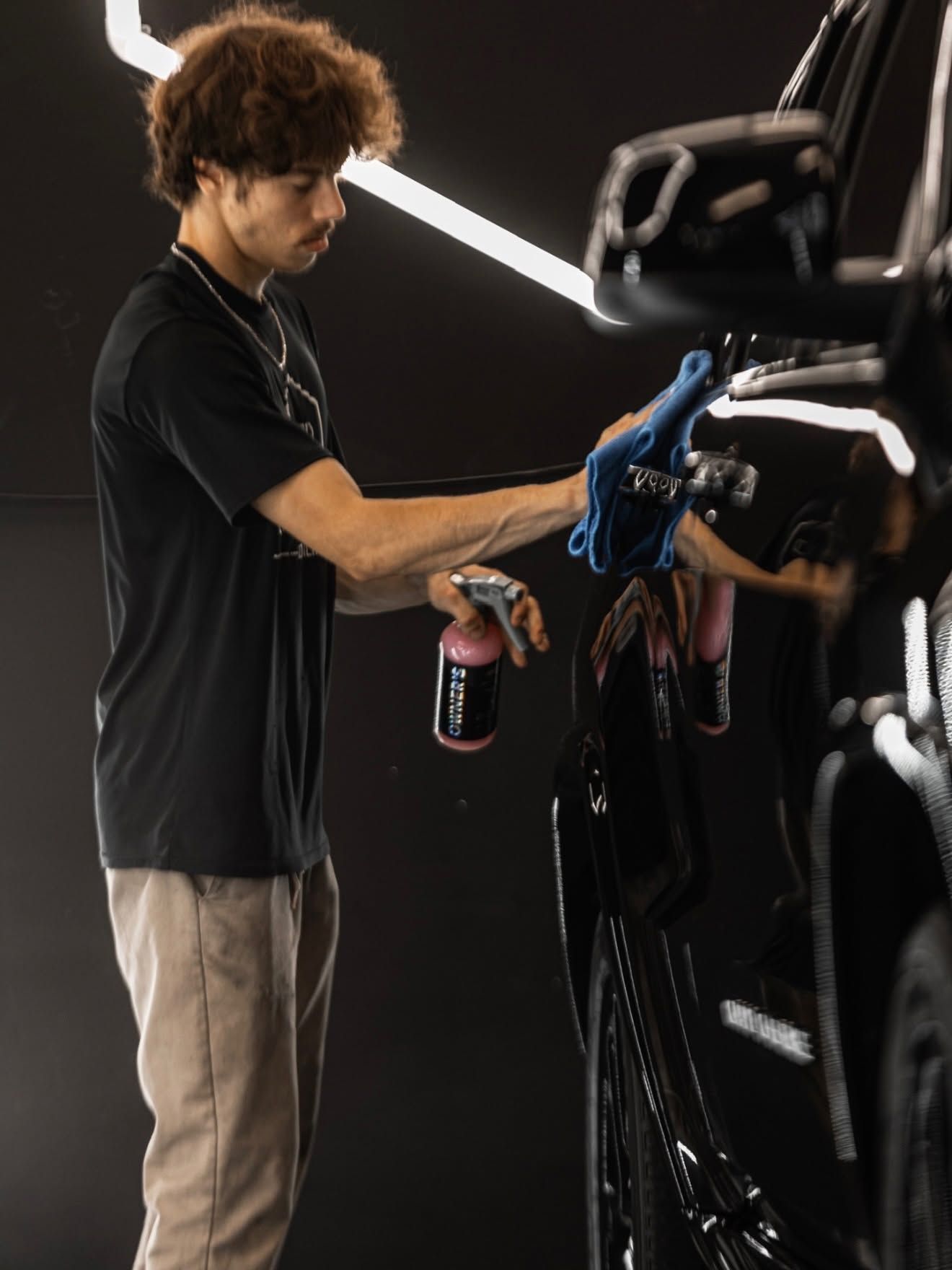 Person detailing a black car with spray bottles in a dark studio with bright diagonal lights