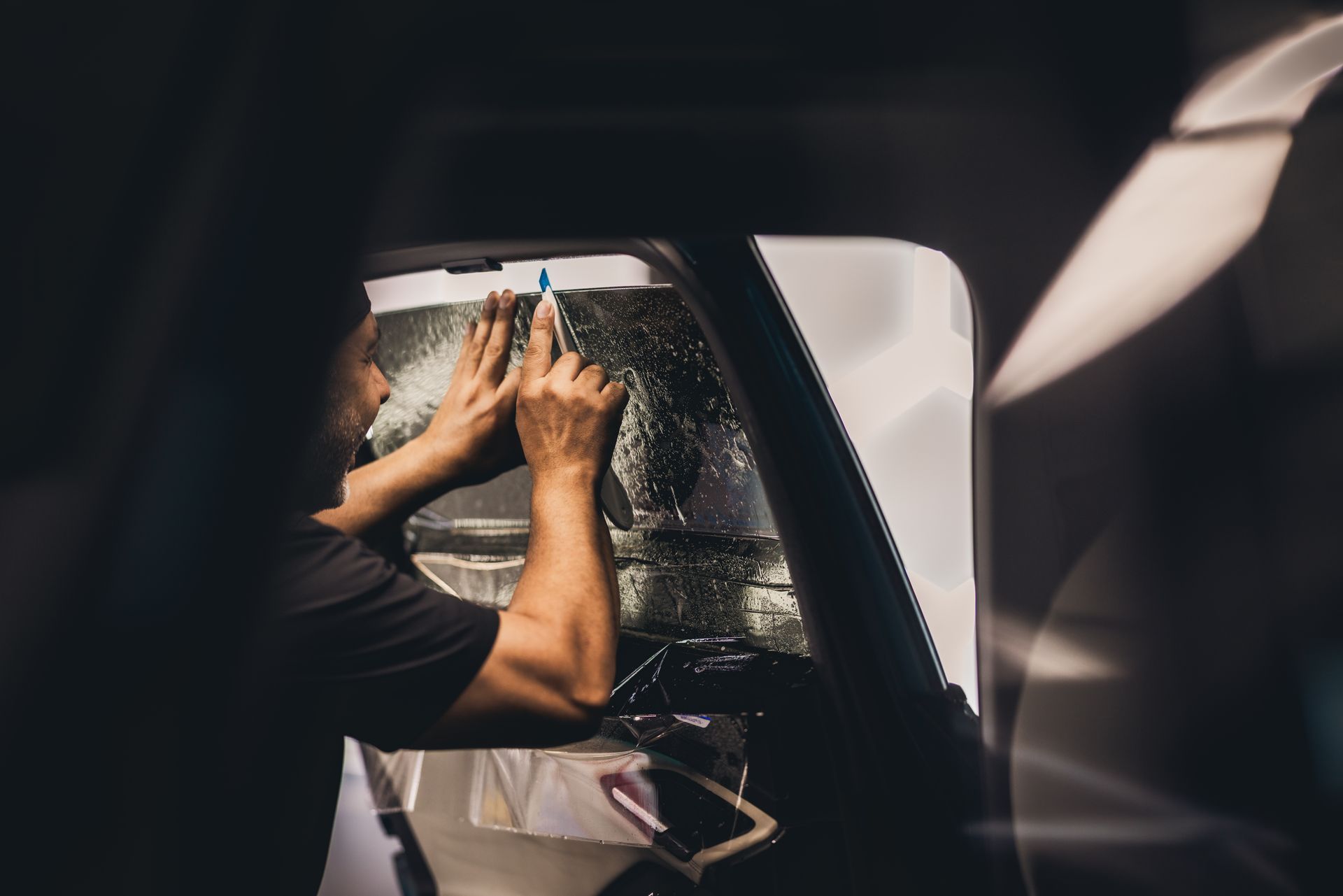 Hands wiping a wet car window from inside the vehicle, with sunlight and reflections outside.