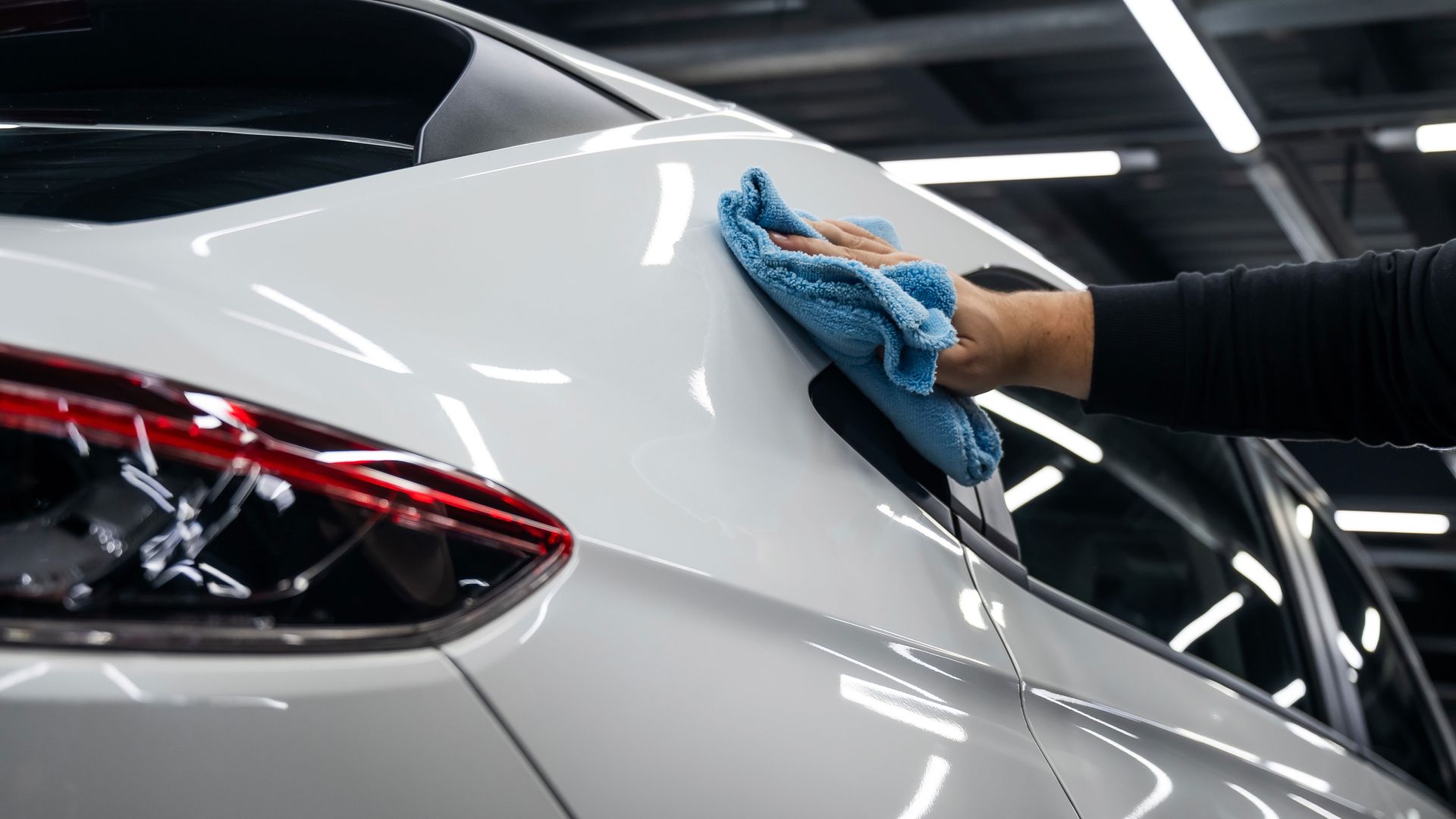 Person wiping the rear of a silver car with a blue cloth in a garage