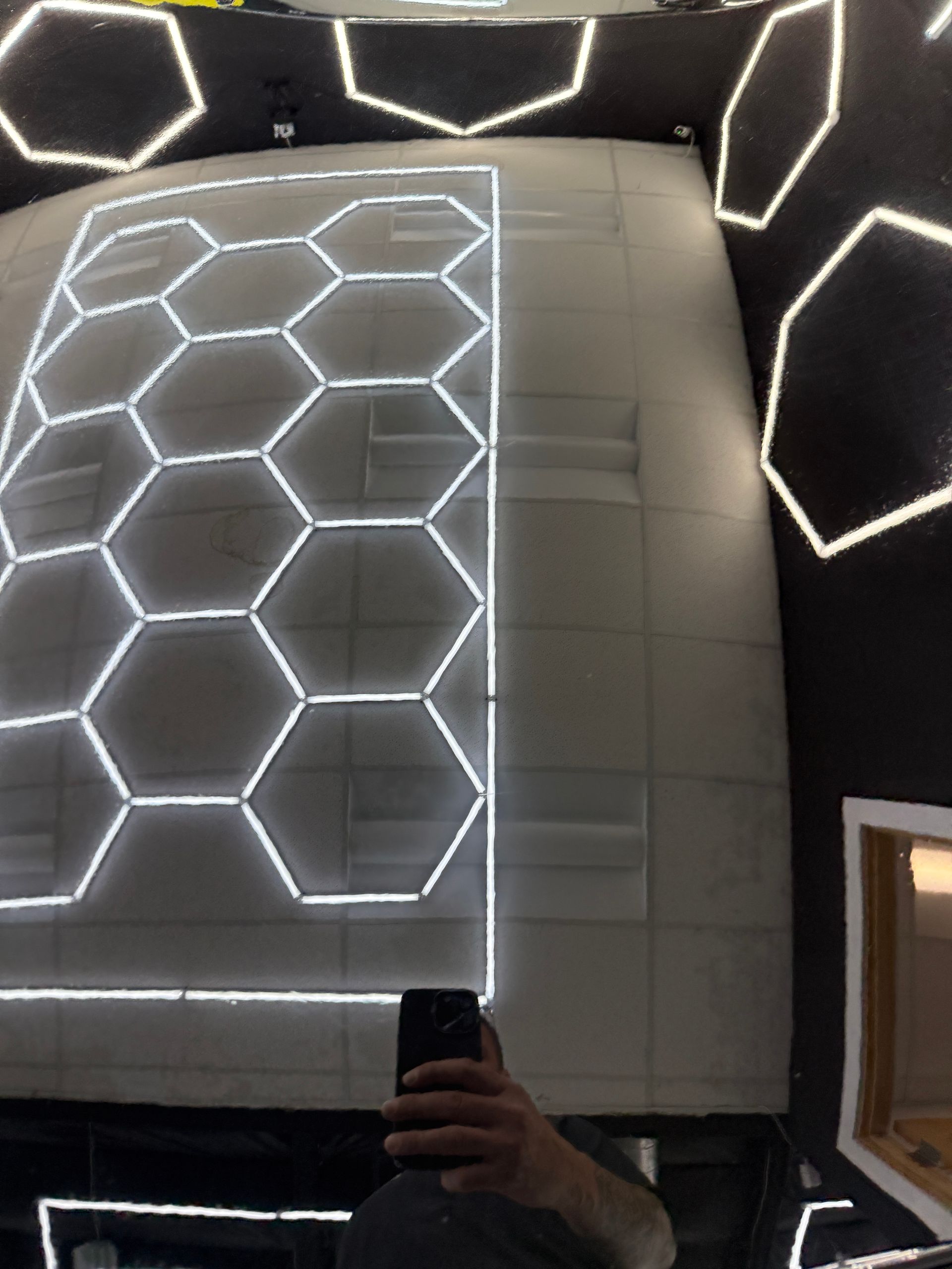 Person taking a mirror selfie in a room with glowing hexagonal wall lights and a curved reflective surface.