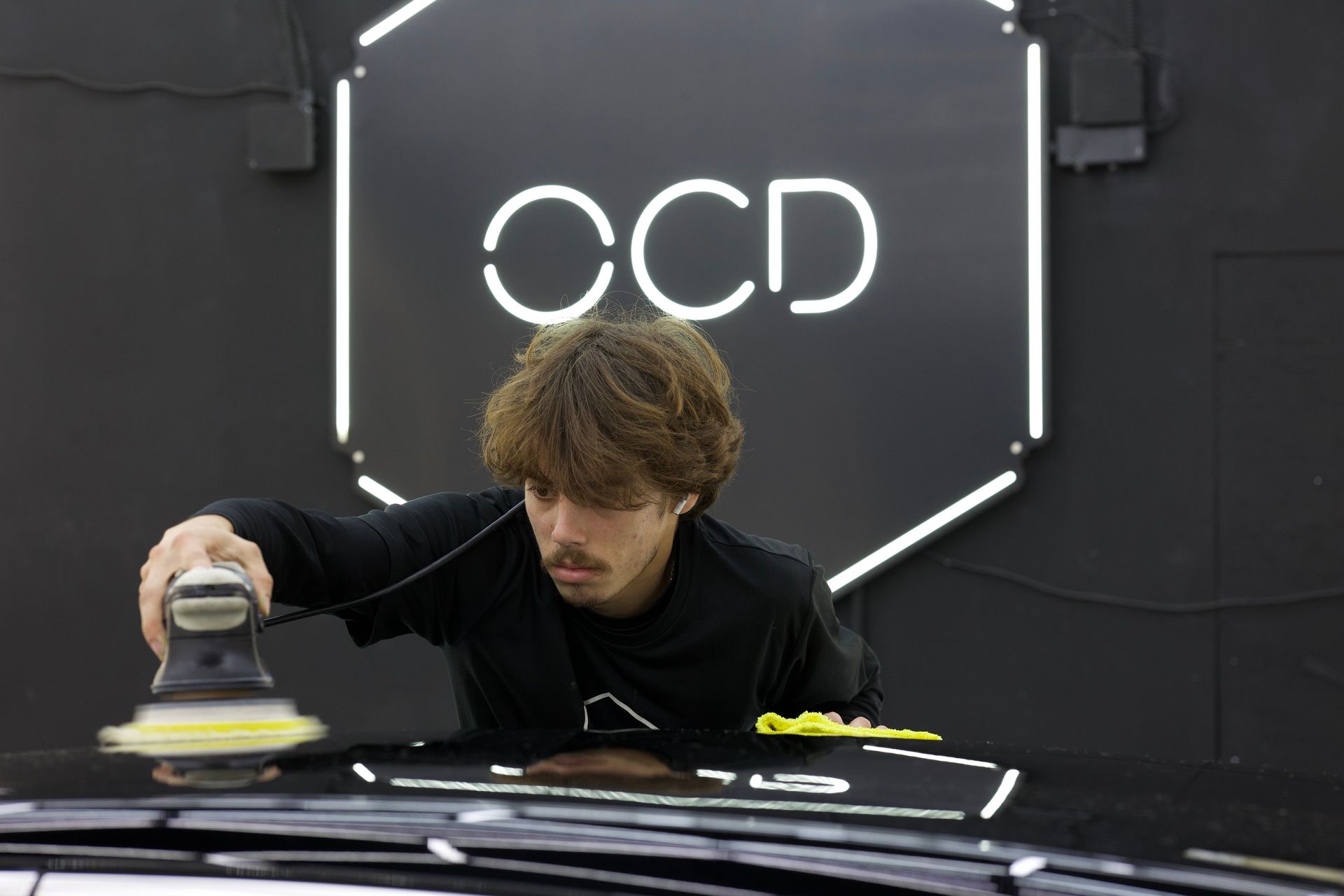 Person polishing a black car in a dark workshop with a glowing CCD sign in the background
