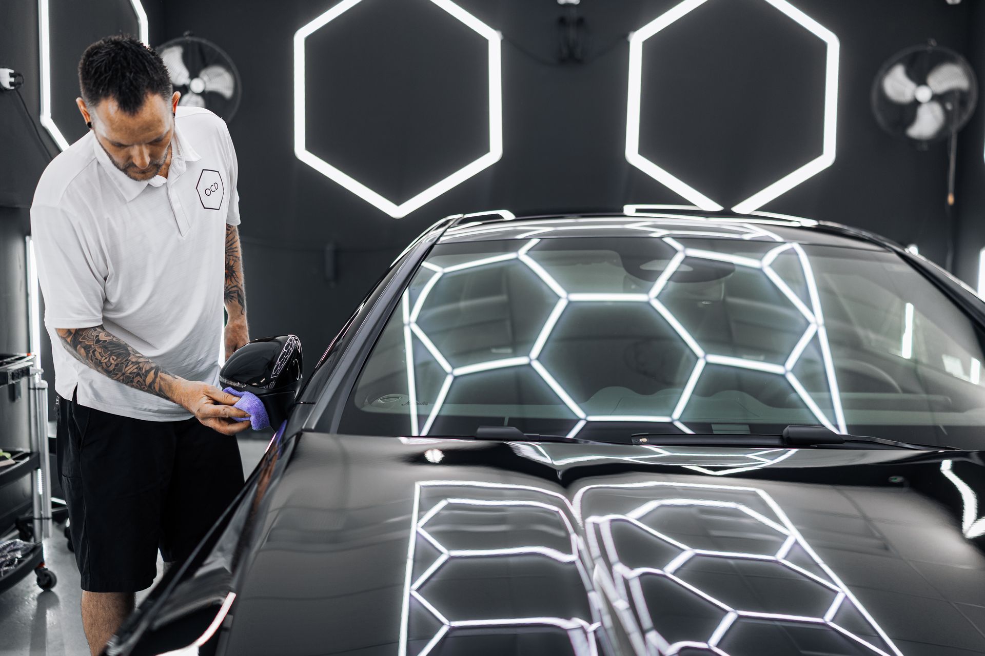 Technician polishing a black car under bright hexagonal lights in a detailing studio
