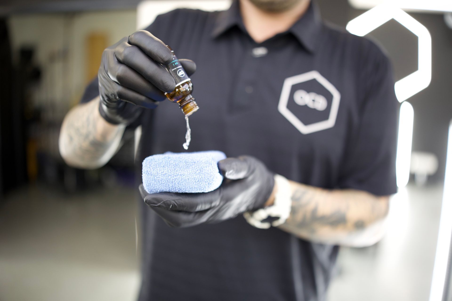 Gloved person applying liquid to a blue microfiber cloth in a garage-like workspace
