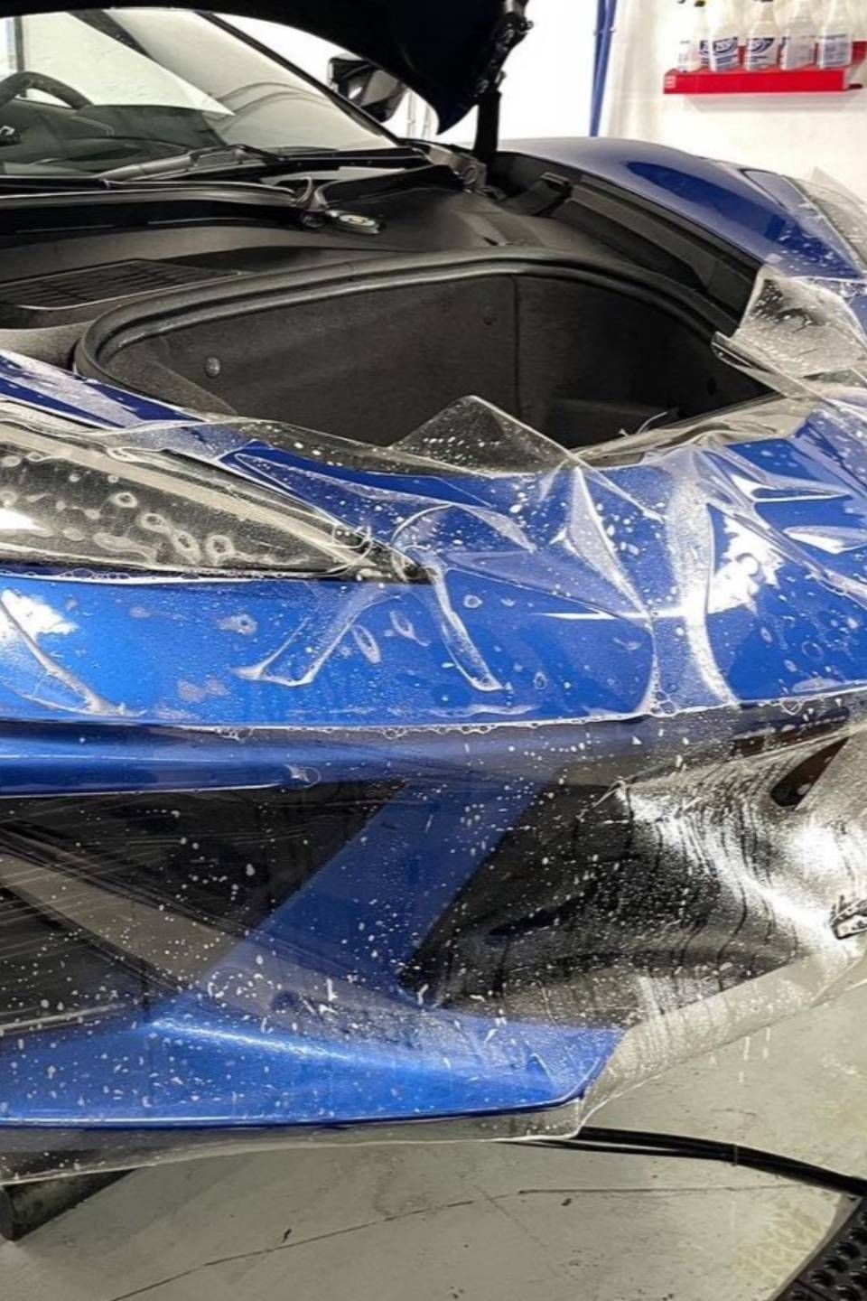 Blue sports car with its front end damaged and hood open, wrapped in protective plastic.
