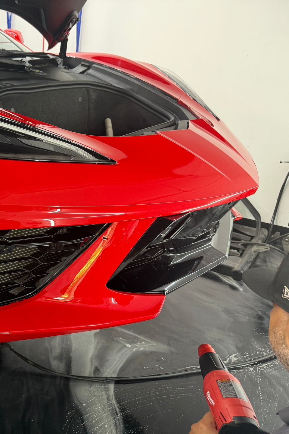 Red sports car front bumper being polished with a handheld buffer in a garage