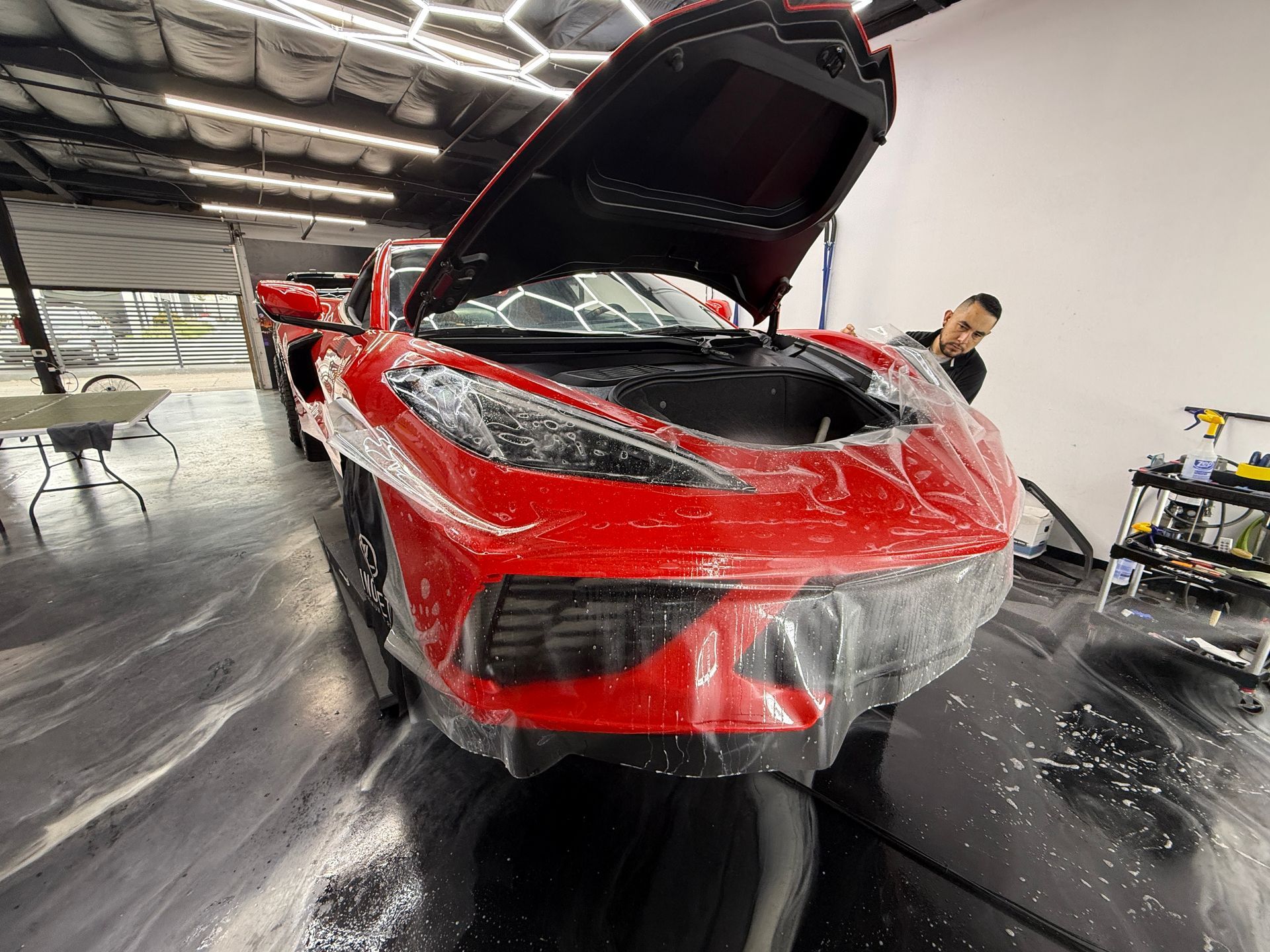 Red sports car wrapped in clear plastic inside a workshop, hood open with a person nearby.