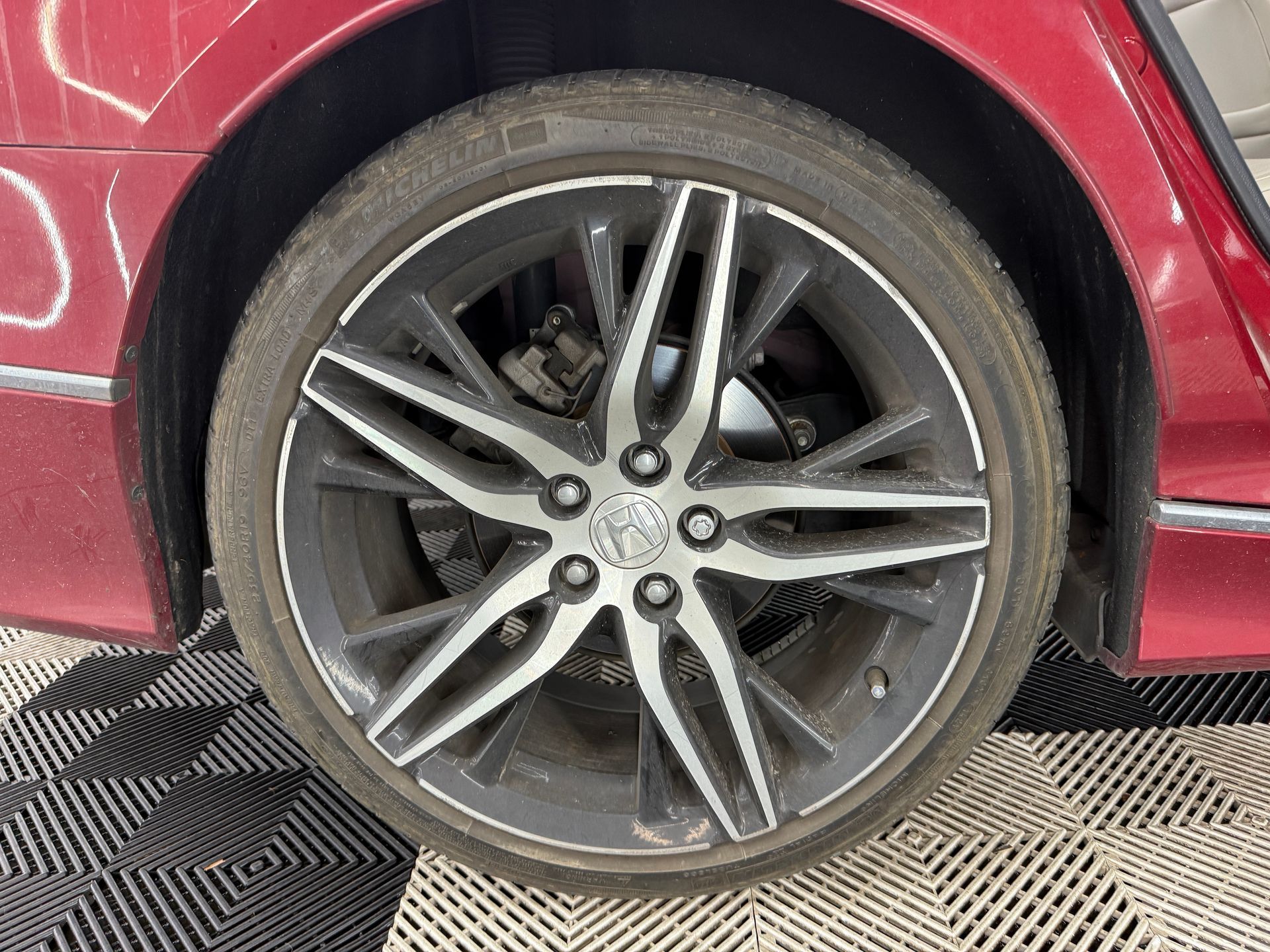 Close-up of a red car’s rear wheel with a silver alloy rim, parked on a patterned floor.