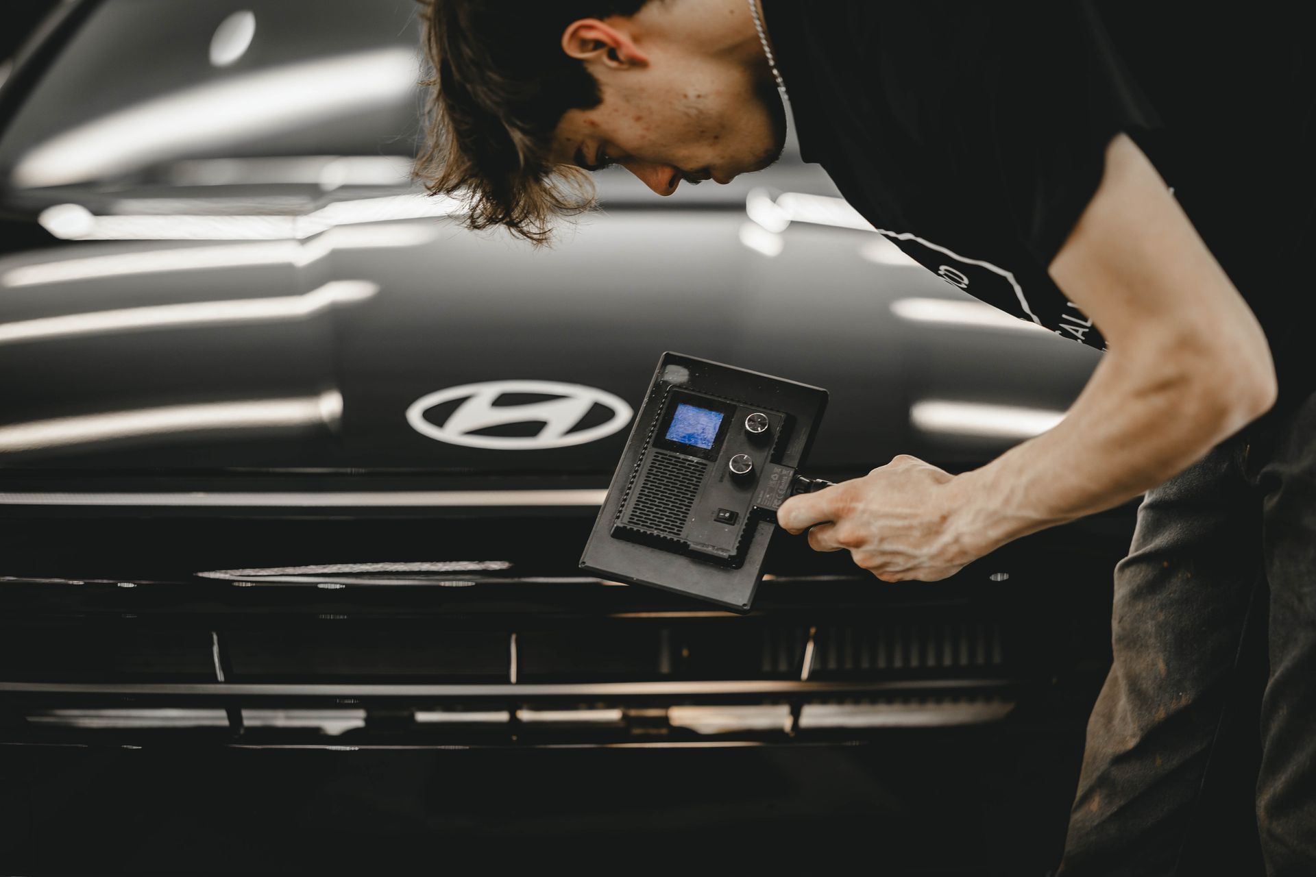 Technician inspecting the front of a black Hyundai with a handheld device in a garage