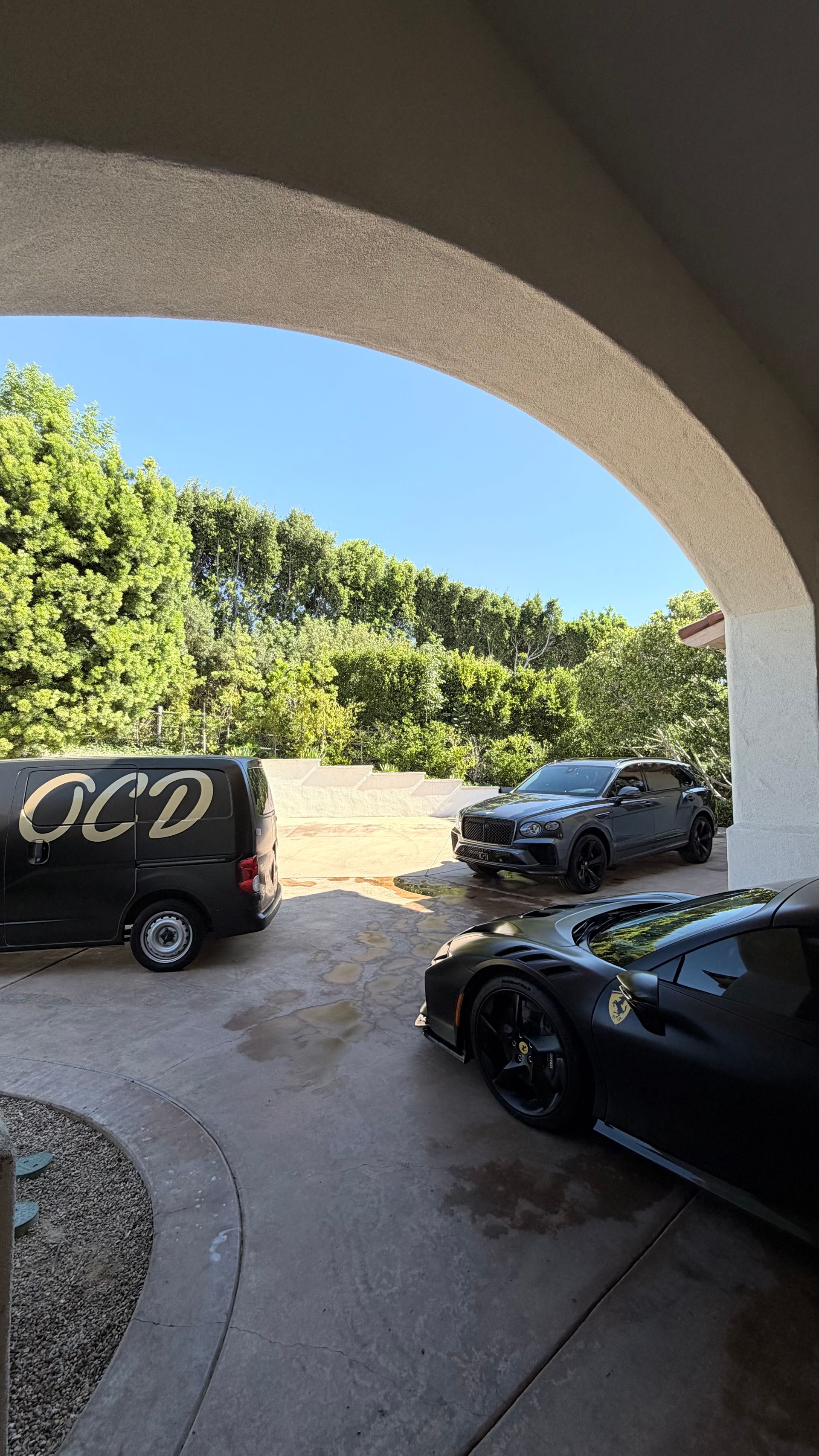 Driveway under an archway with two parked cars and a black van, bordered by tall green hedges.