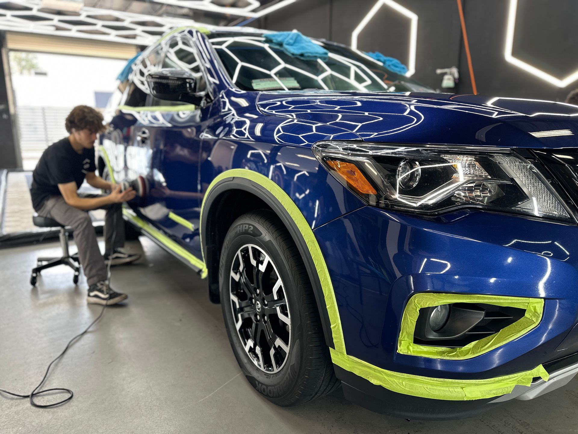 Worker detailing a blue SUV in a garage, with neon green masking tape around the trim.