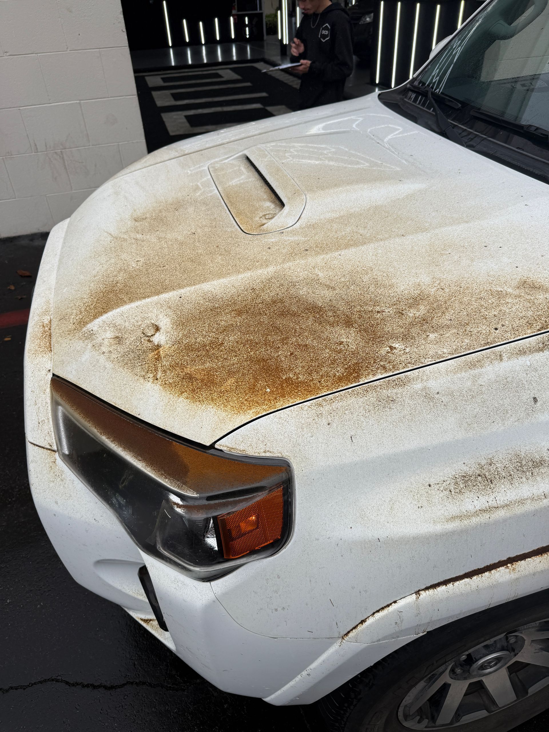 White SUV hood and front fender covered in orange-brown rust speckles in a driveway