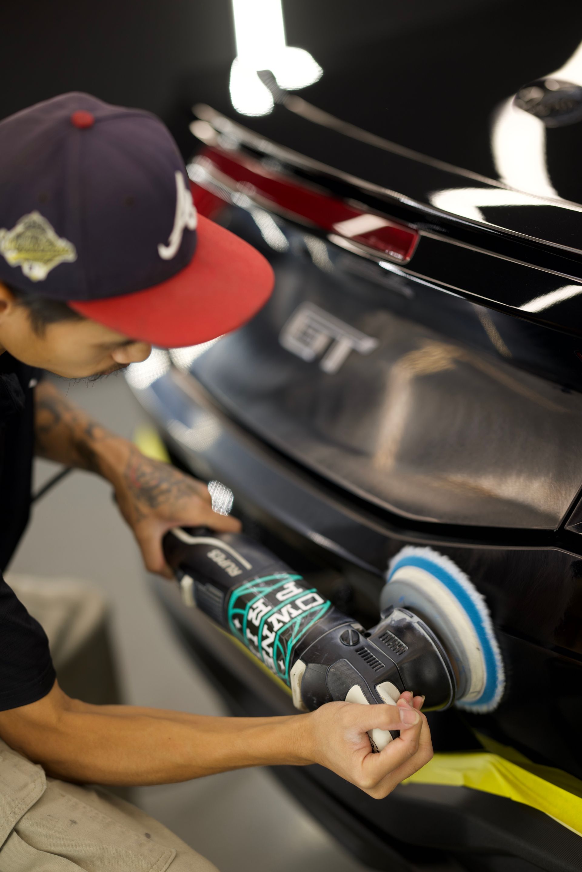 Person polishing a black car’s rear bumper with a power buffer in a workshop