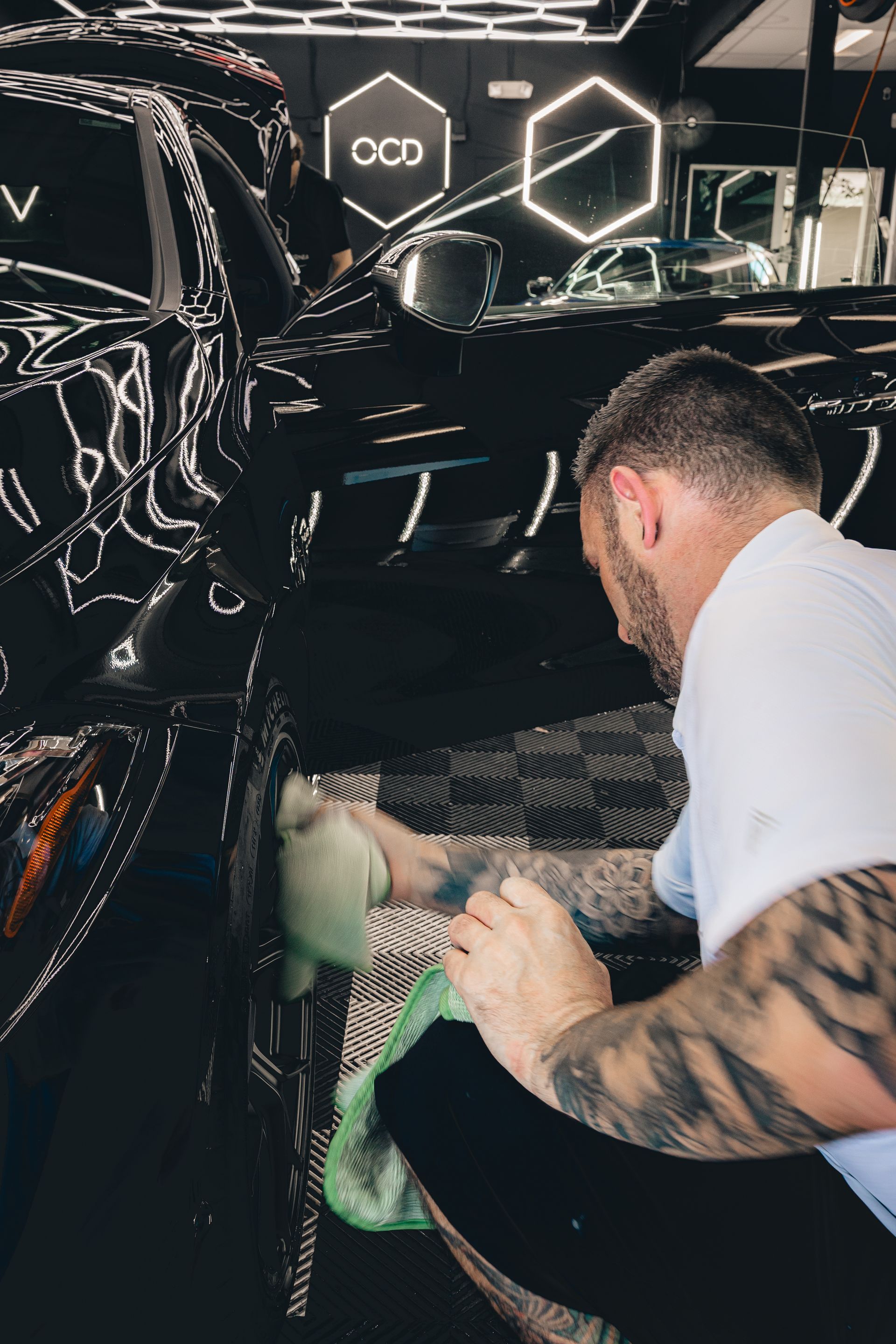 Man polishing a black car in a garage with illuminated hexagon lights in the background