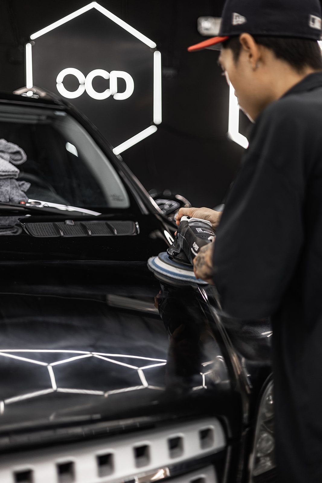 Person detailing a black car in a shop with a glowing OCD sign in the background