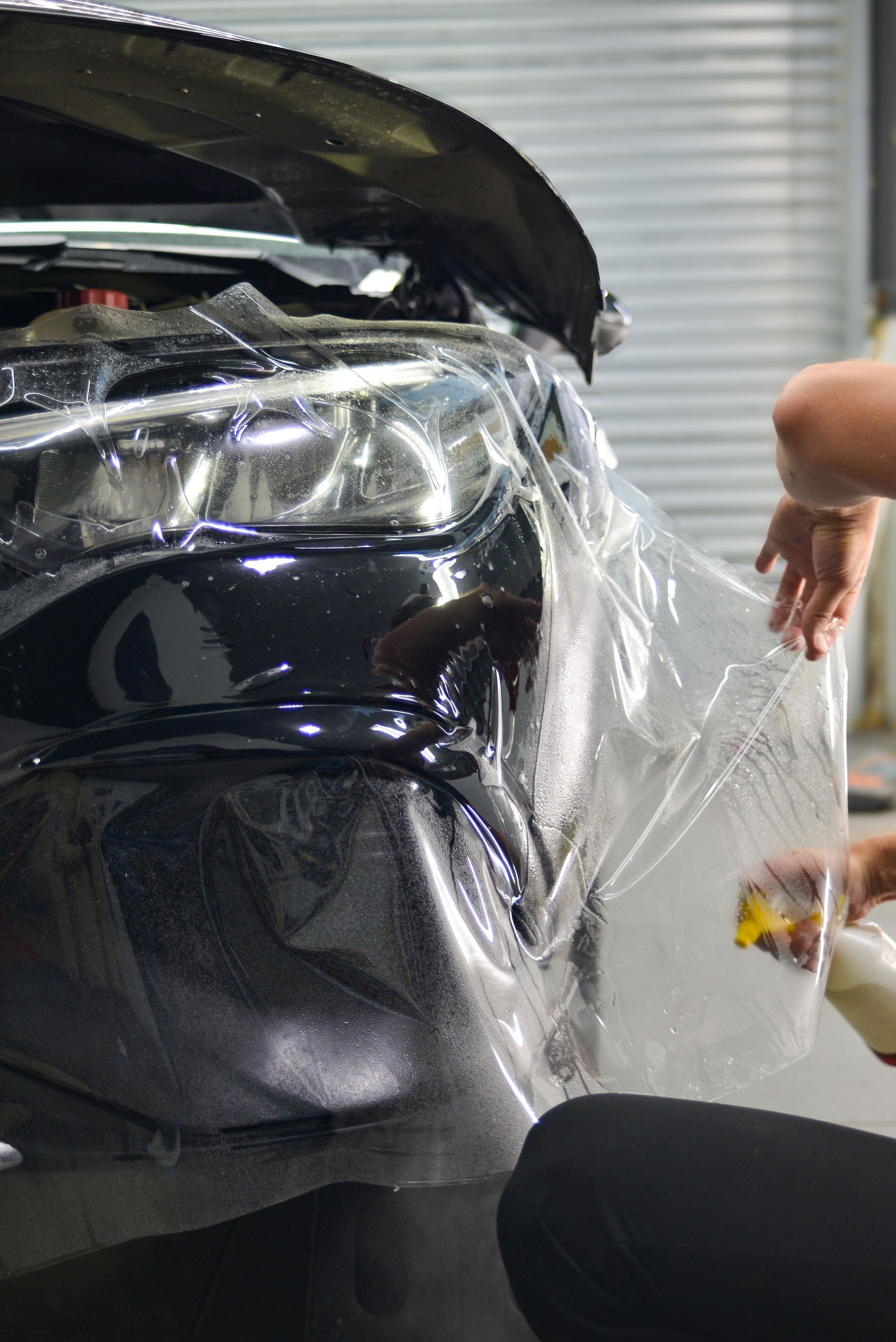 A black car’s front end wrapped in clear protective film, with a hand reaching beside it.