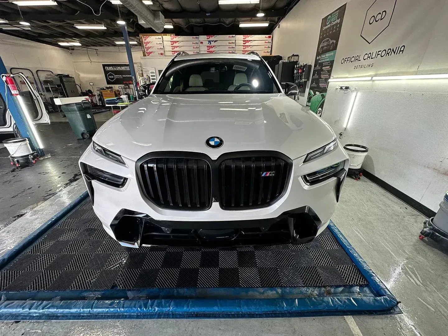 White BMW SUV on a lift in an auto repair shop, front view with black grille and trim.